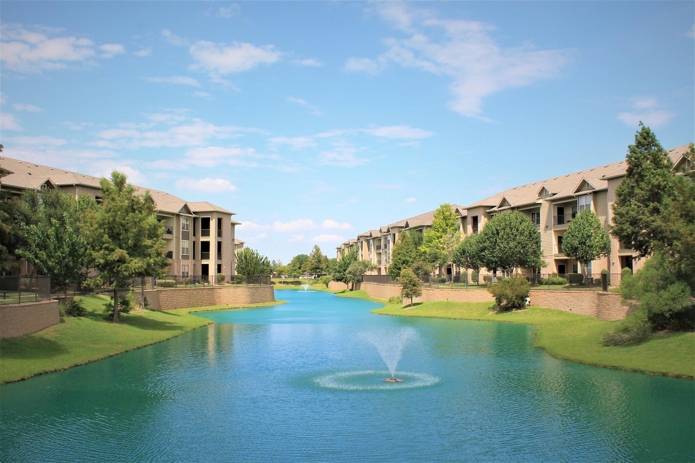 A serene view of a calm green river running between residential buildings. Trees line the banks, and a small fountain creates ripples in the water under a clear blue sky with scattered clouds. The scene captures a peaceful suburban setting.