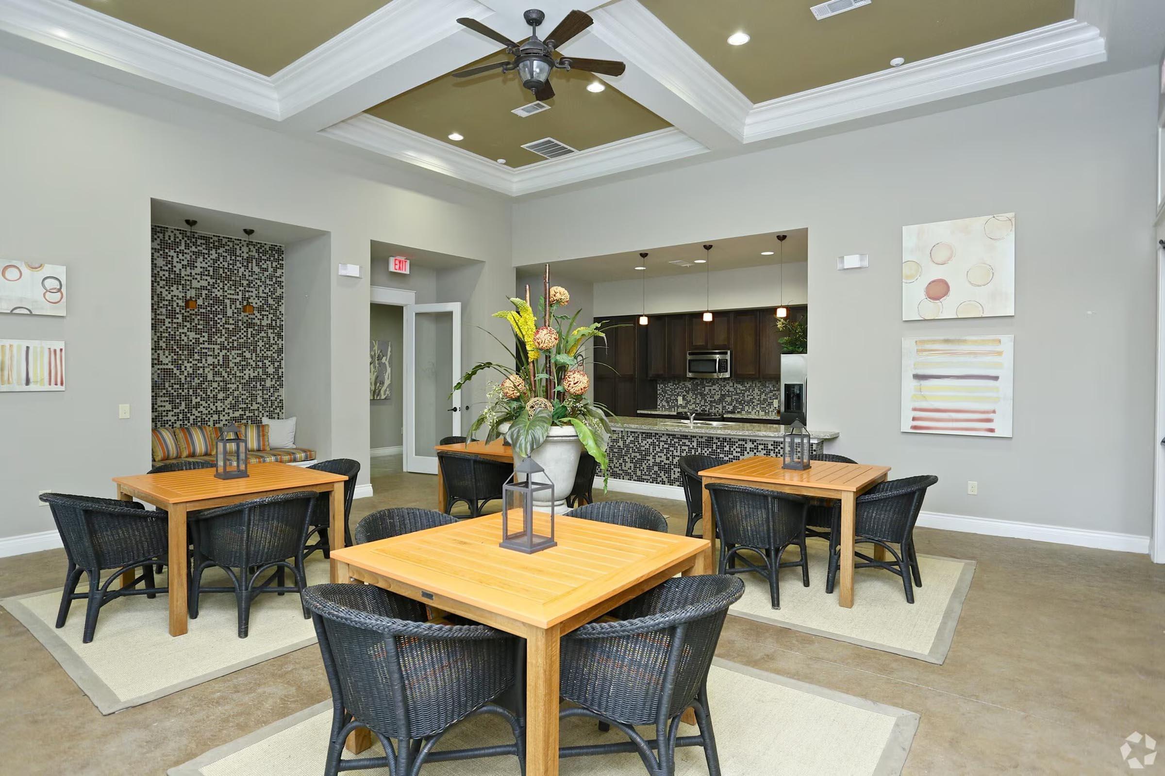 A modern community lounge featuring several wooden tables surrounded by black wicker chairs. The room has light gray walls, a decorative accent wall with patterns, and large windows. A large potted plant serves as a centerpiece on one table, and there are framed artworks on the walls.