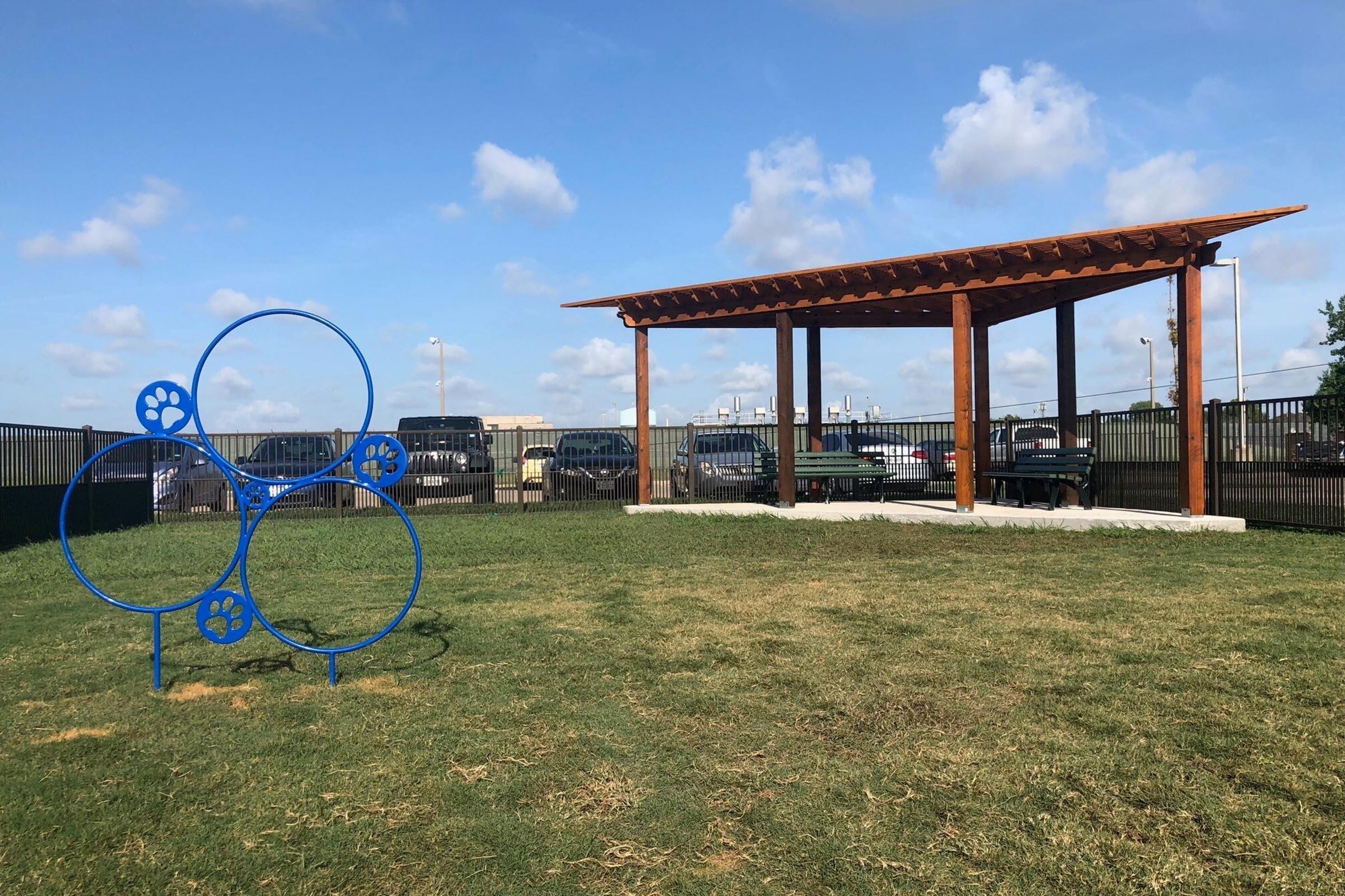 A spacious outdoor dog park featuring a grassy area, a wooden pavilion with a slatted roof, and a blue dog-themed play structure shaped like overlapping circles with paw prints. The park is enclosed by a black fence and features clear blue skies with a few clouds in the background.