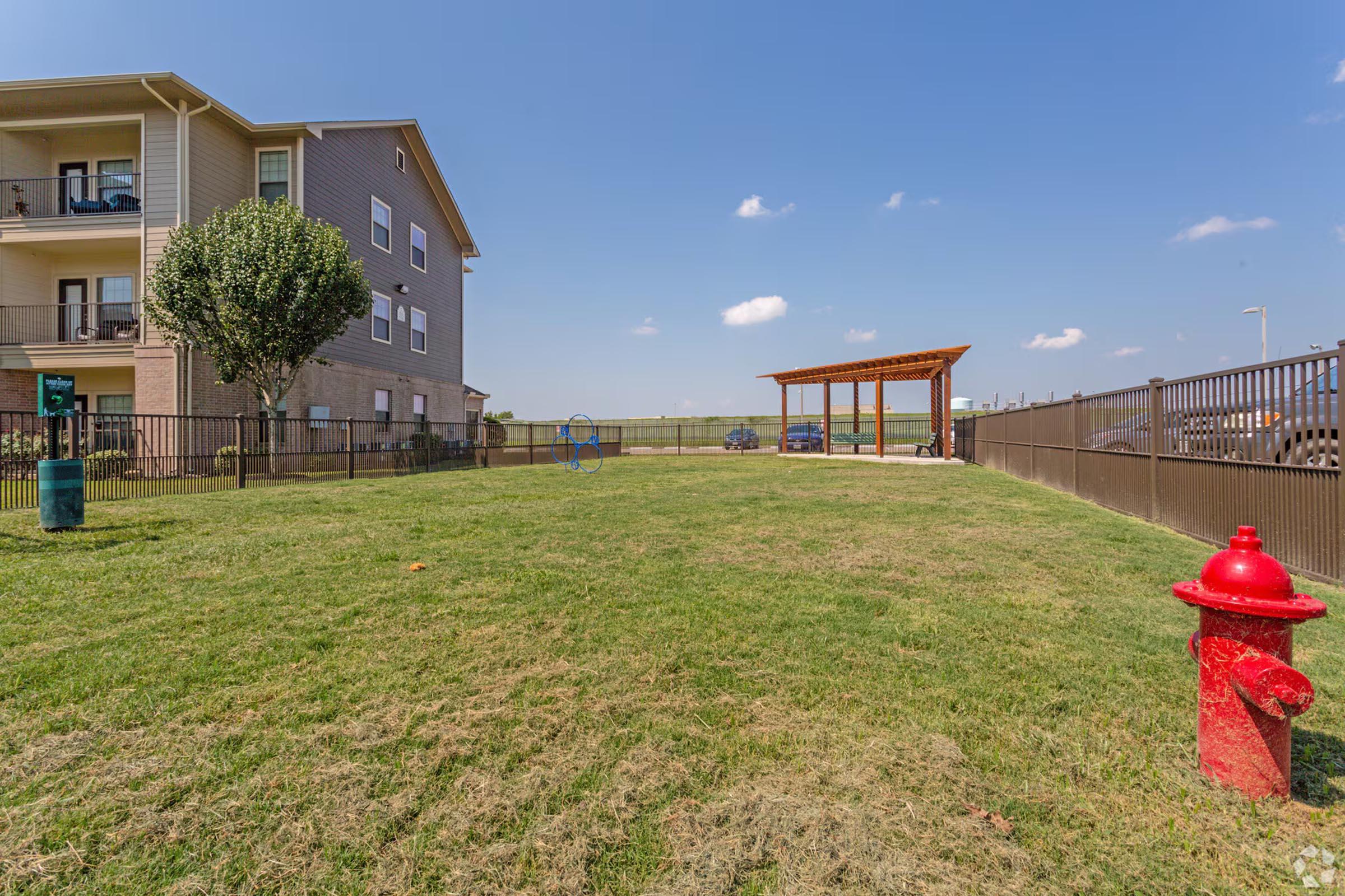 A grassy area in a residential complex featuring a red fire hydrant, a wooden gazebo structure, and a fence. In the background, there are a few parked cars and an apartment building. The sky is clear with a few scattered clouds.