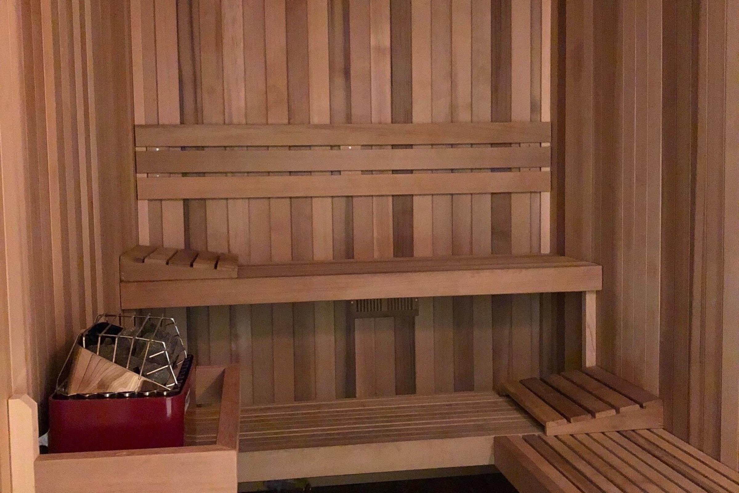 Interior of a wooden sauna featuring wooden benches, with a small corner for sauna stones and a heater. The walls and benches are made of light-colored wood, creating a warm and inviting atmosphere.