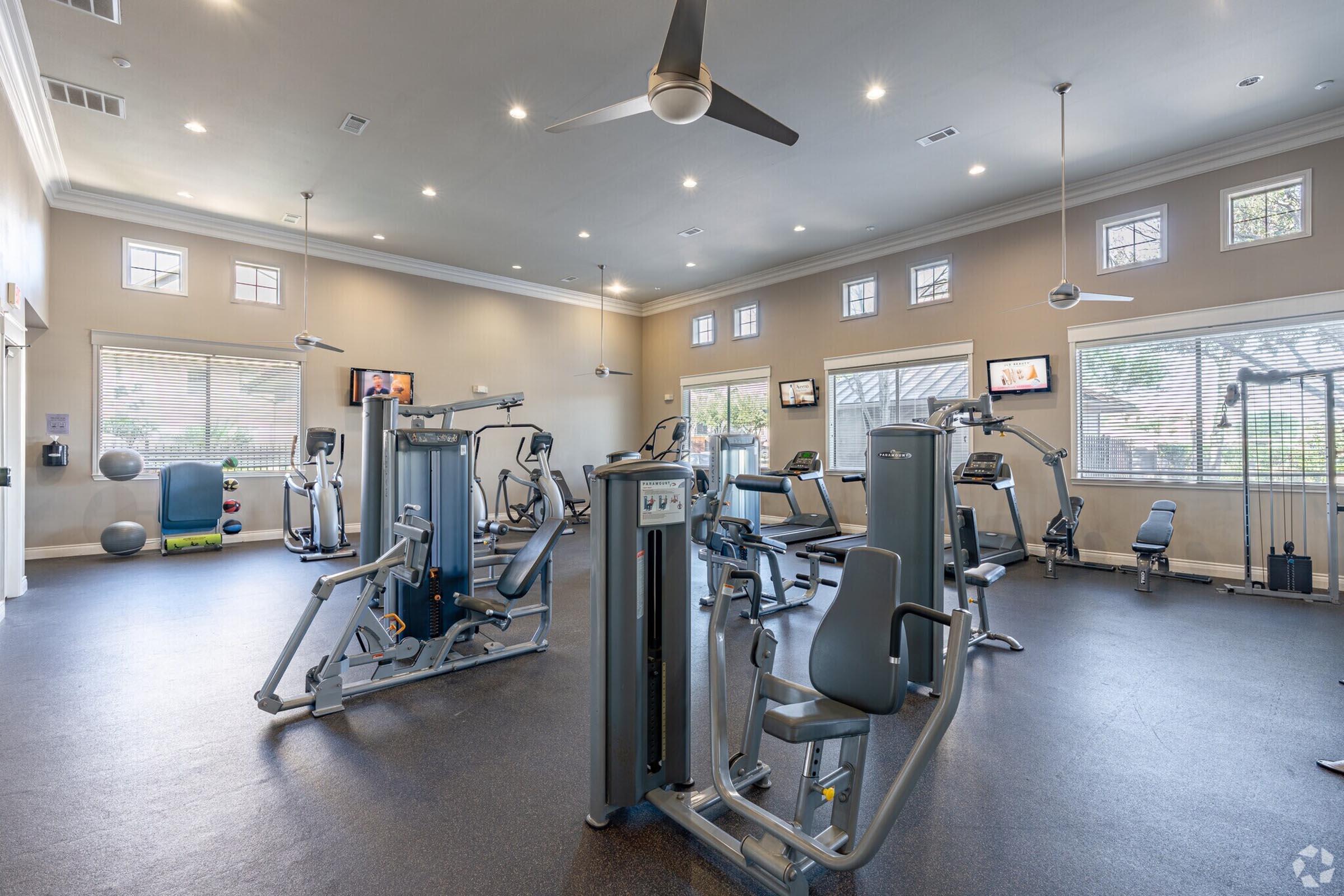 A modern gym interior featuring various workout machines, including weight machines and cardio equipment. The space is well-lit with natural light from large windows, and flat-screen TVs are mounted on the walls. There are exercise balls and a ceiling fan for ventilation.
