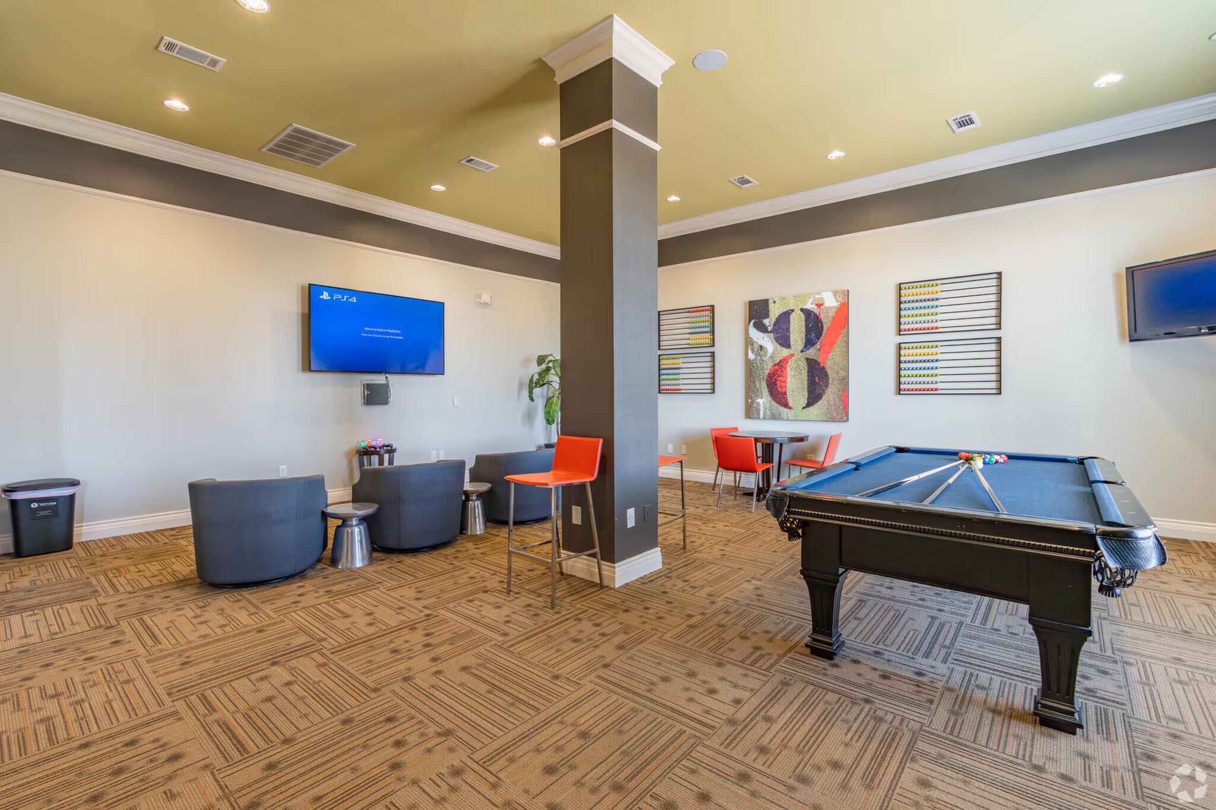 Modern lounge area featuring a pool table, seating arrangements with gray chairs, a television mounted on the wall, and a colorful abstract painting. The space also includes a dining table with orange chairs, creating a vibrant and inviting atmosphere.