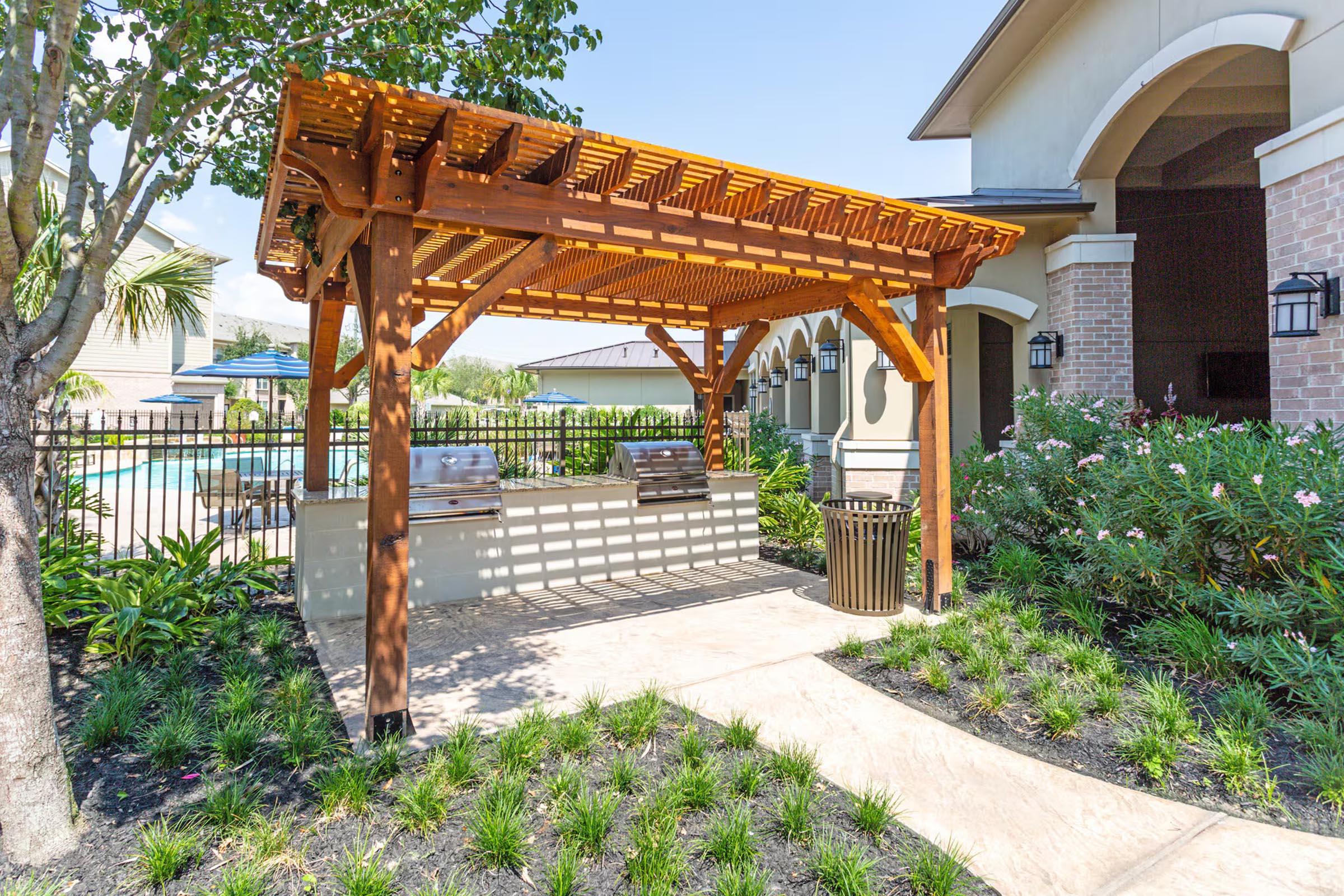A wooden pergola with seating underneath, surrounded by landscaped greenery, leads to a pool area. The scene features a paved walkway, a trash bin, and a sunny sky.