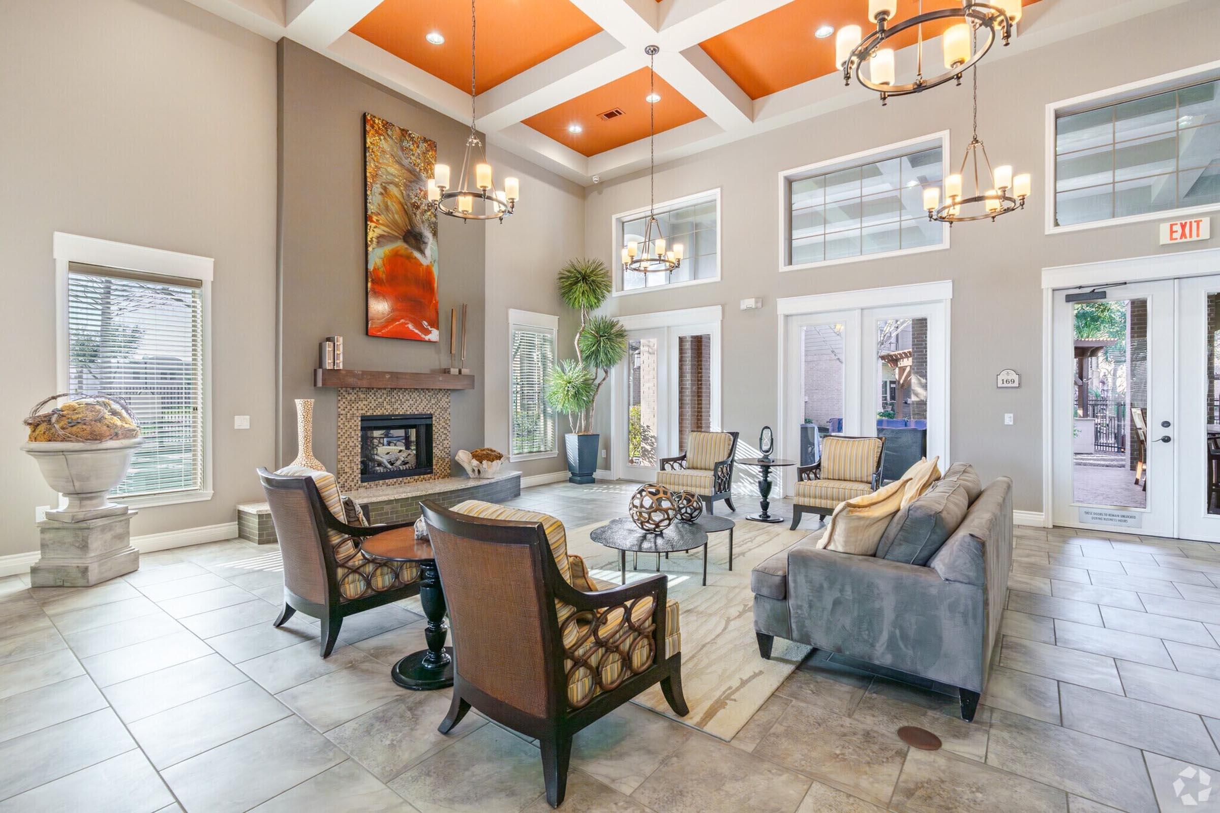 A spacious and modern lounge area featuring large windows, a stylish fireplace, and contemporary furniture. The ceiling is adorned with orange accents, and there are various indoor plants. The room is well-lit with chandelier lighting, creating an inviting atmosphere.