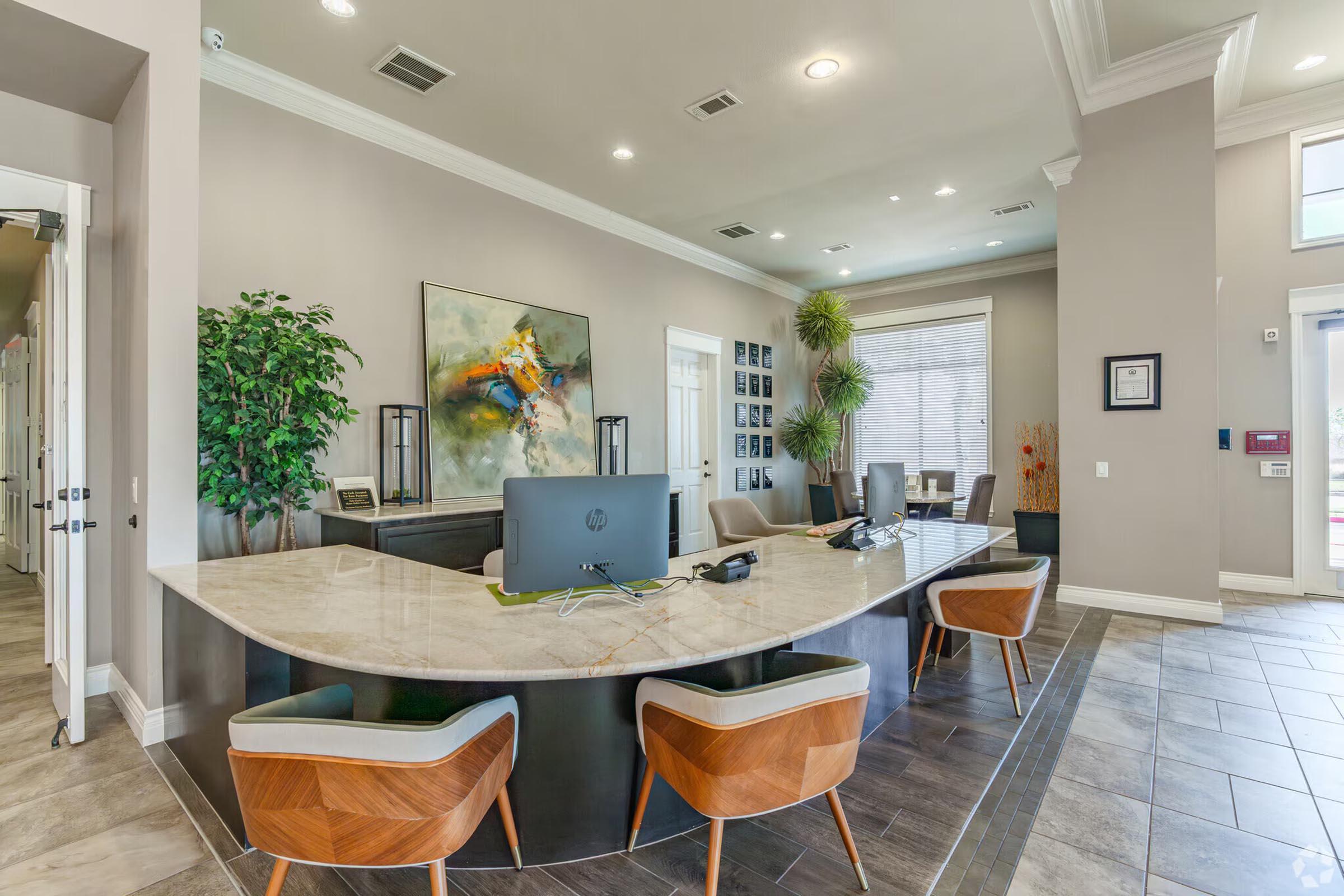 Modern reception area featuring a curved marble counter with a computer, stylish wooden chairs, and decorative plants. The walls are painted in neutral tones, complemented by artwork and a series of framed photos. Ample lighting creates a welcoming atmosphere.