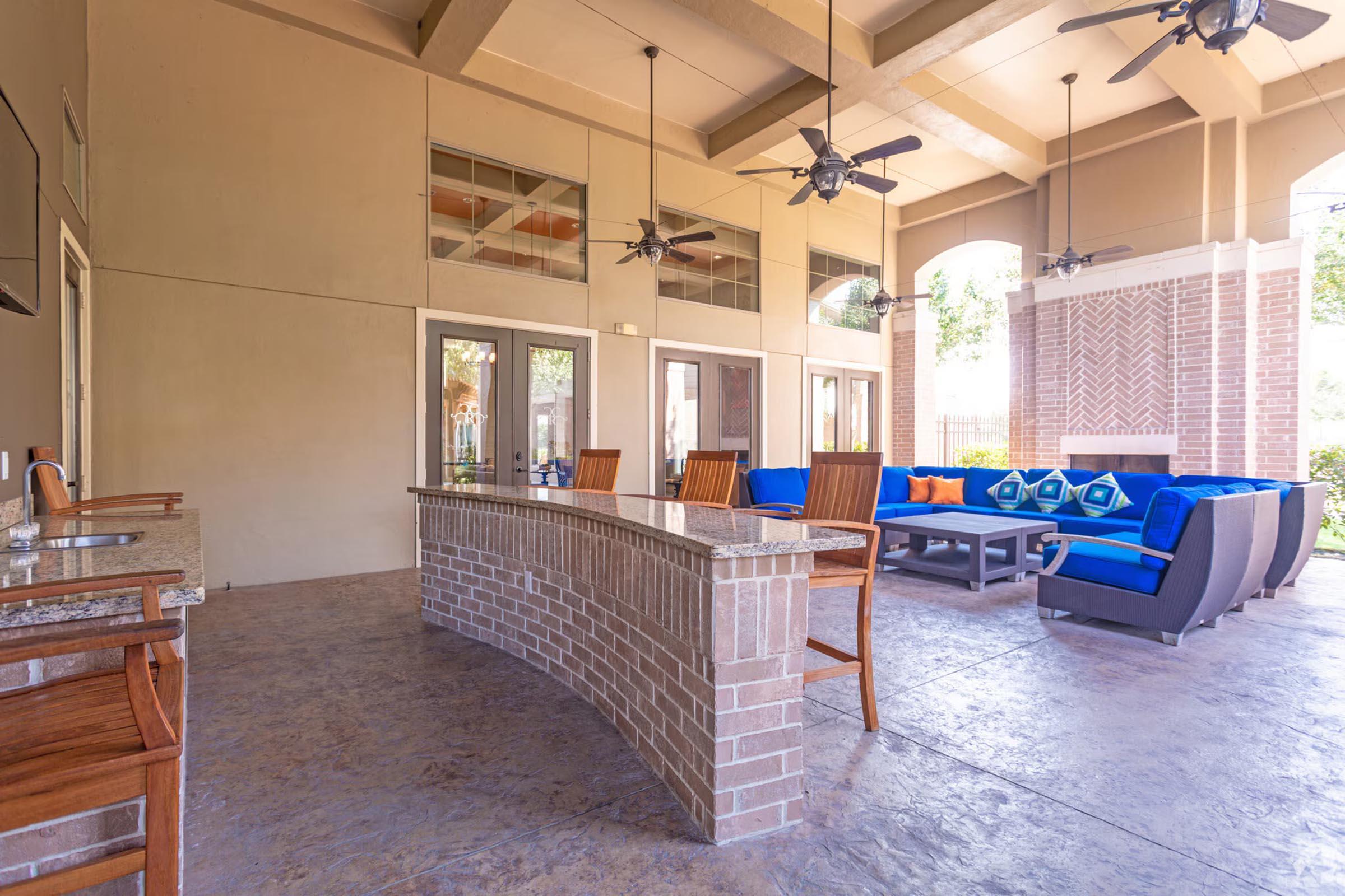 Spacious outdoor patio area featuring a bar with high stools, comfortable blue and orange seating, and ceiling fans. Natural light floods the space through large windows. The flooring is stylishly finished, enhancing the inviting atmosphere for relaxation or gatherings.