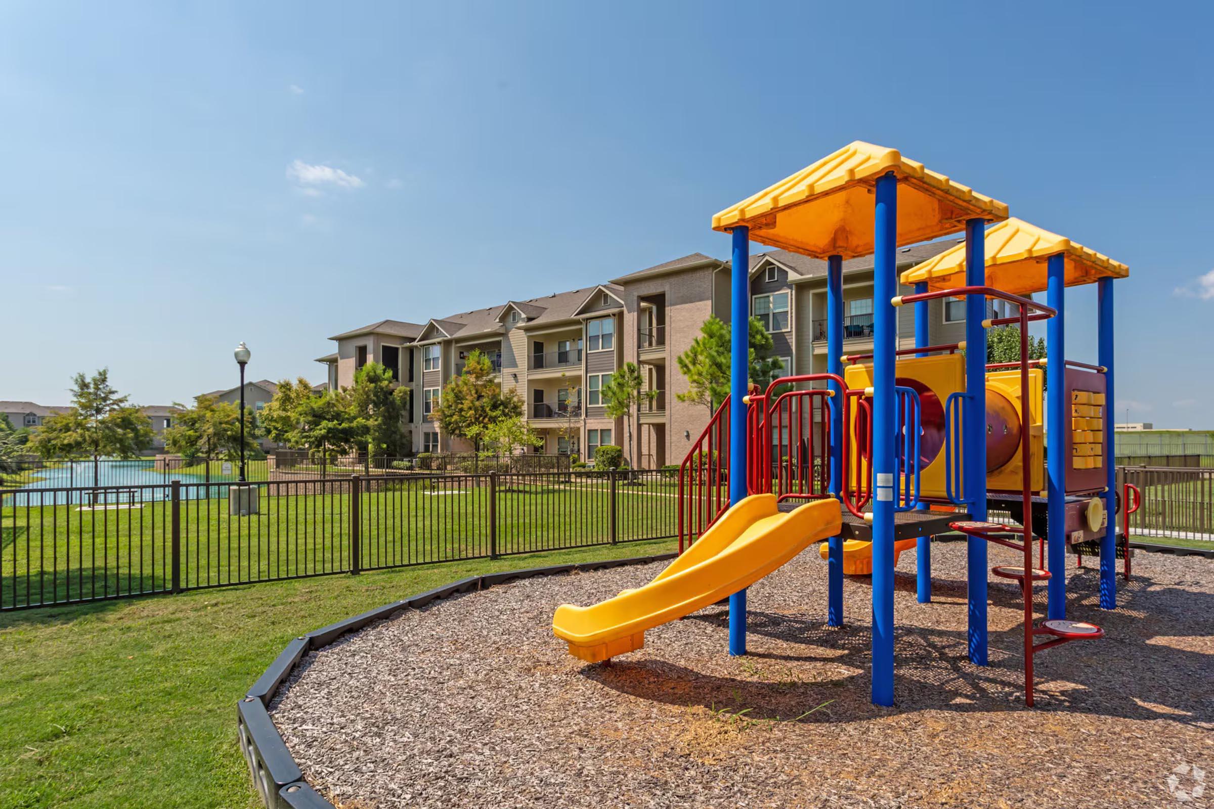 Playground equipment featuring a yellow slide, climbing structures, and colorful posts, set in a landscaped area with an apartment building in the background. The grassy space includes fencing and trees, creating a family-friendly outdoor environment on a sunny day.