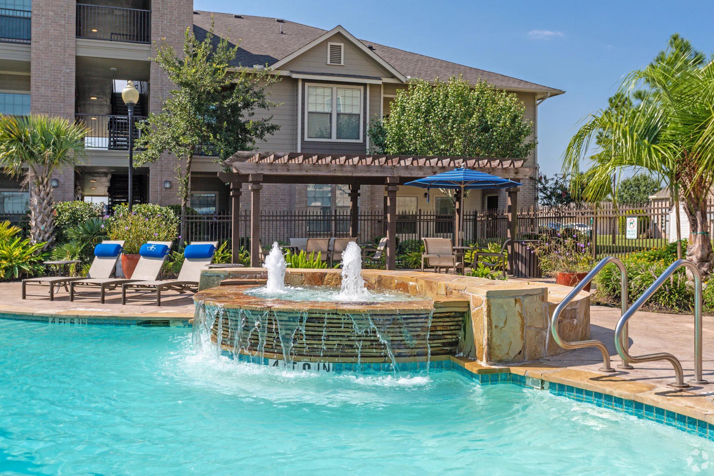A serene outdoor pool area featuring a central fountain, lounge chairs, and landscaped greenery. In the background, an apartment building is visible, along with pergolas and palm trees, creating a relaxing atmosphere for residents. Clear blue skies add to the inviting setting.