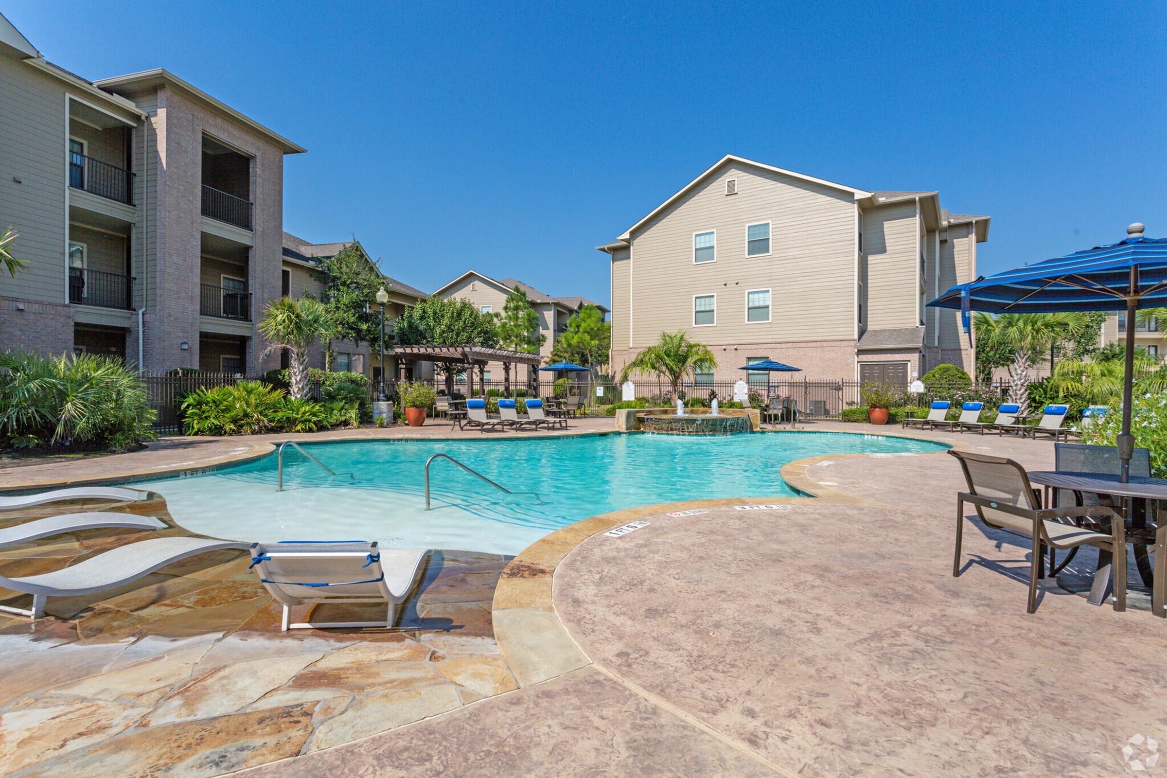 Outdoor swimming pool at an apartment complex, surrounded by lounge chairs and umbrellas. Lush greenery and landscaped areas are visible in the background. Two multi-story buildings with balconies are seen beside the pool, all under a clear blue sky.