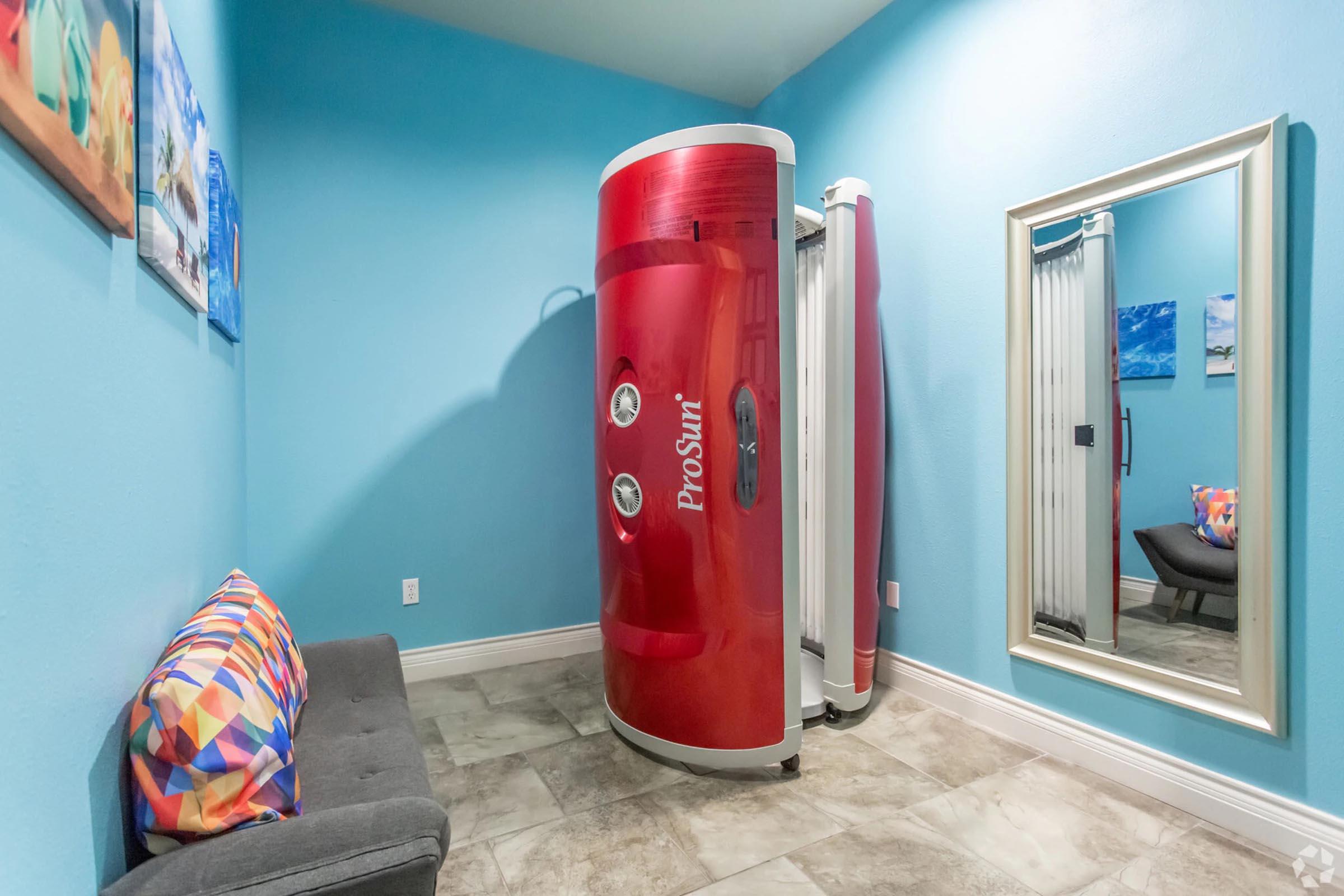 A vibrant room featuring a red tanning booth with a mirror and a gray couch. The walls are painted light blue, adorned with colorful artwork. The flooring is tiled, and the space is well-lit, creating a welcoming atmosphere for tanning.