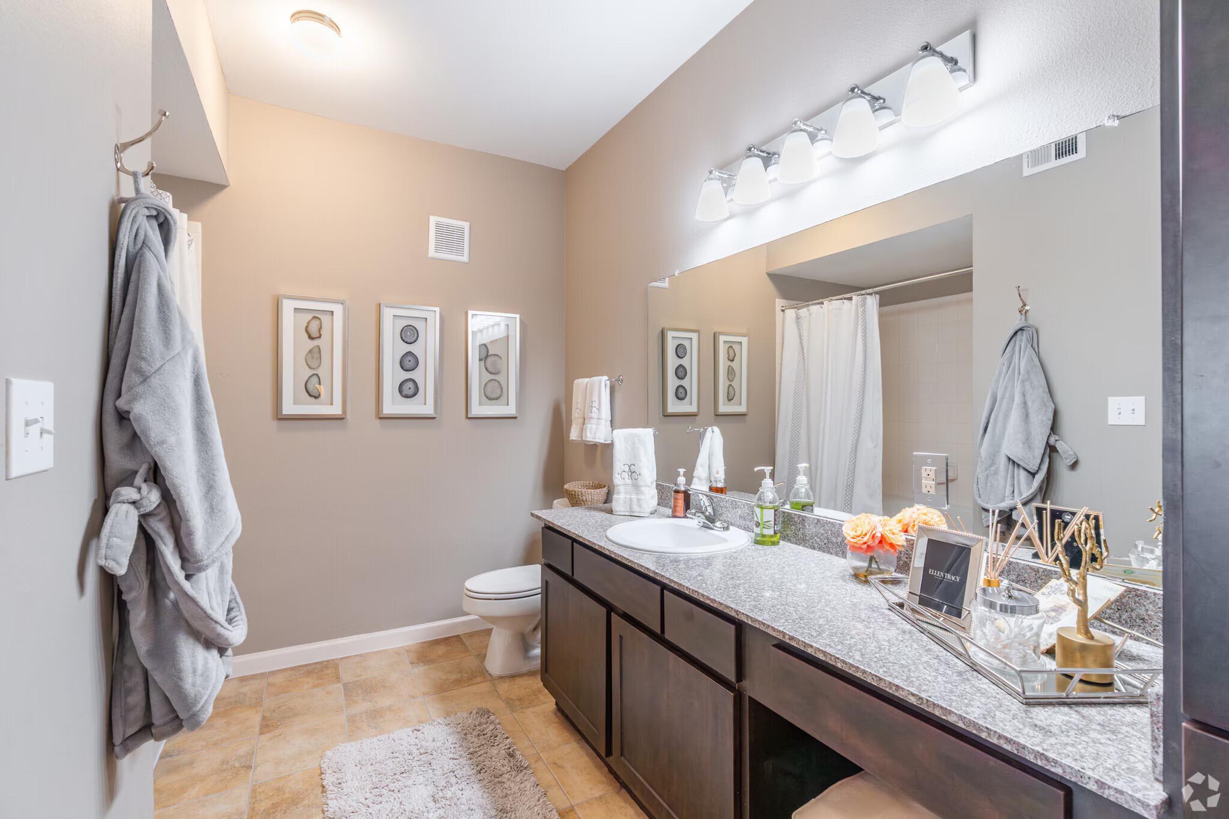 Modern bathroom featuring a double vanity with a granite countertop, two framed wall art pieces, and neatly hung towels. A shower curtain and beige floor tiles complete the space, along with decorative items and a small bouquet of flowers. The lighting is bright and inviting.