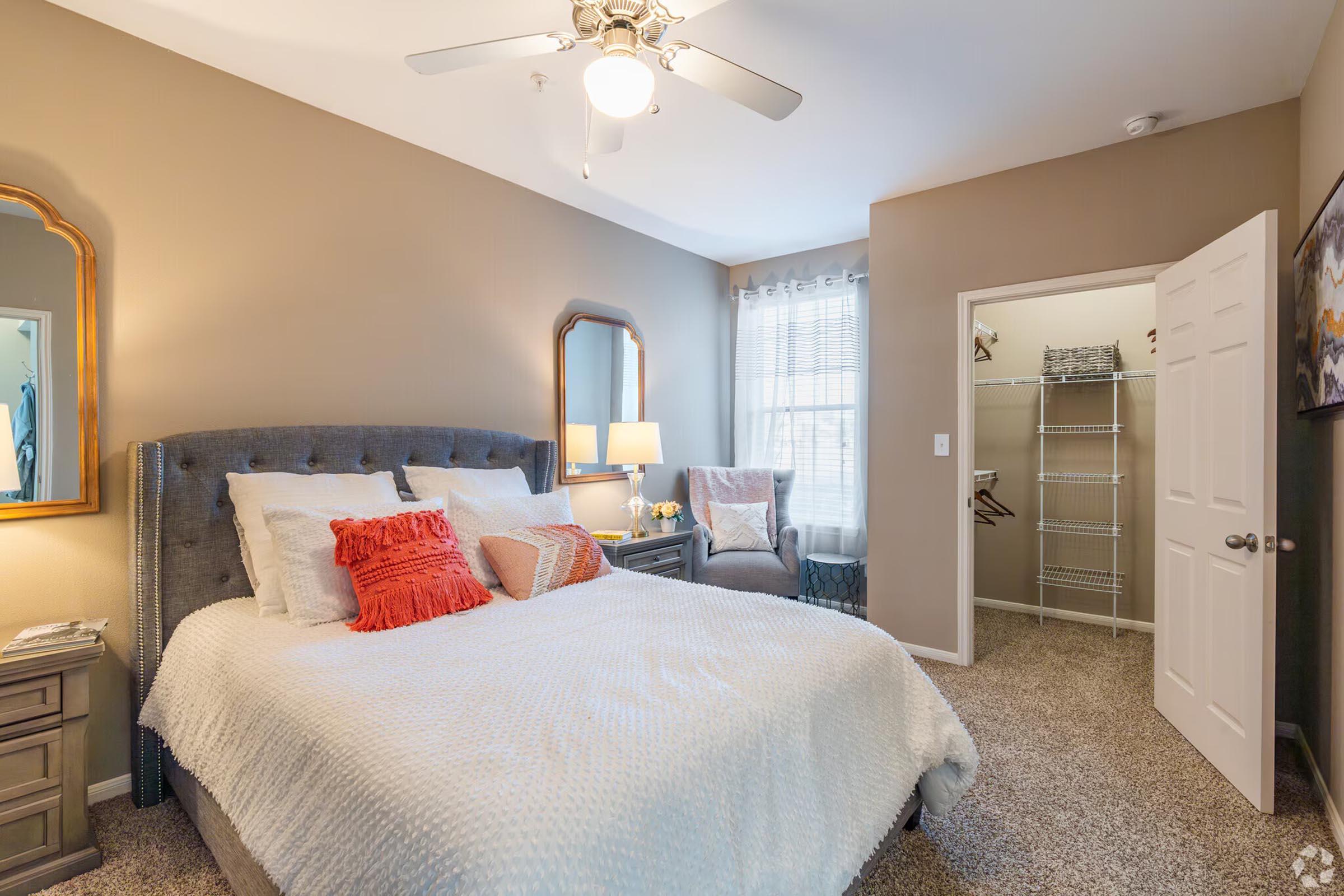 Cozy bedroom featuring a plush bed with decorative pillows, a bedside table with a lamp, and a seating area with a chair. Two mirrors are on the wall, and a closet with shelving is visible. Soft beige walls and a light carpet enhance the warm atmosphere. Natural light filters through the window.