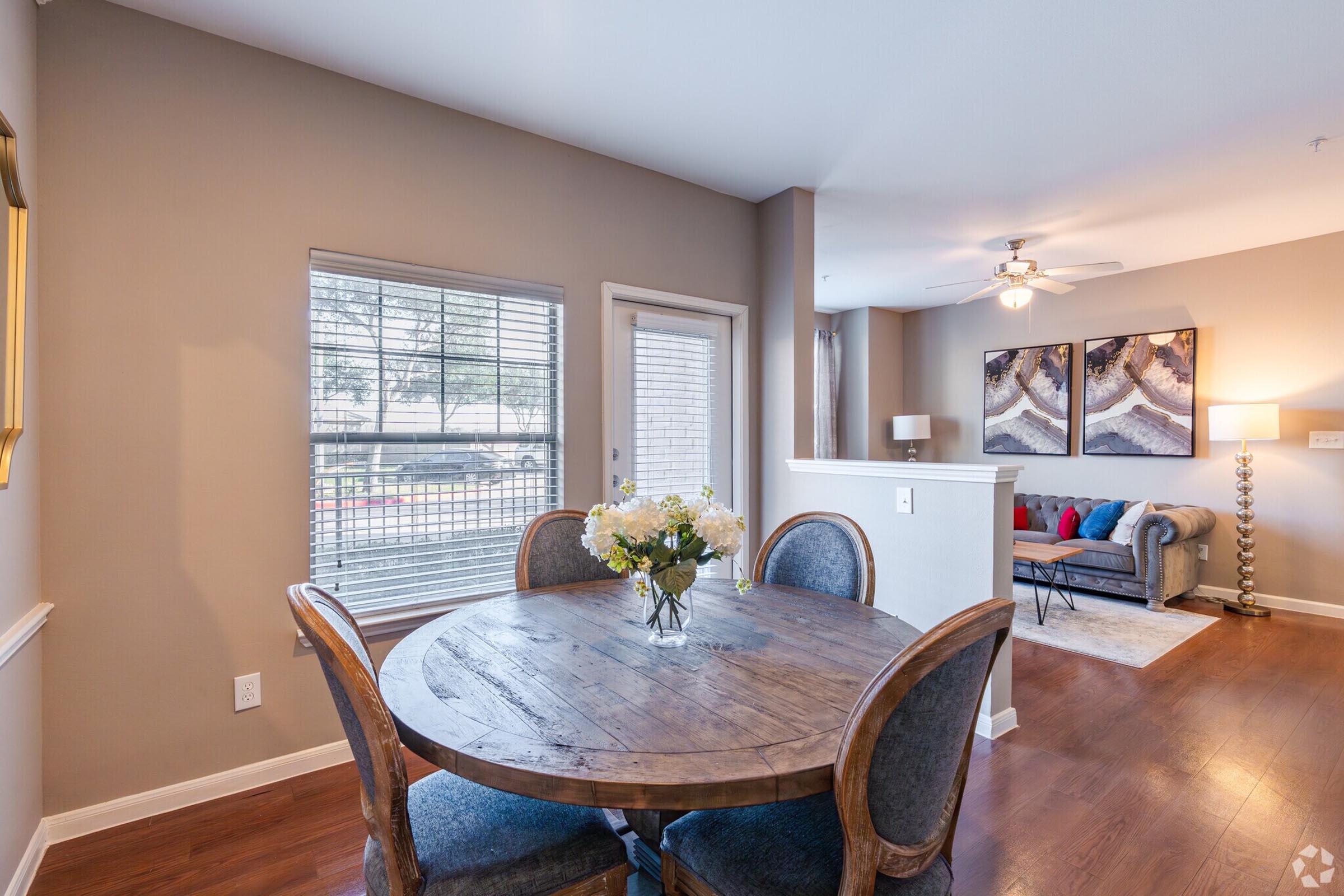 A cozy dining area featuring a round wooden table with blue upholstered chairs, a vase of flowers in the center, and a large window allowing natural light. In the background, there's a comfortable living room with a couch, decorative pillows, and a ceiling fan. Soft neutral tones create a warm atmosphere.