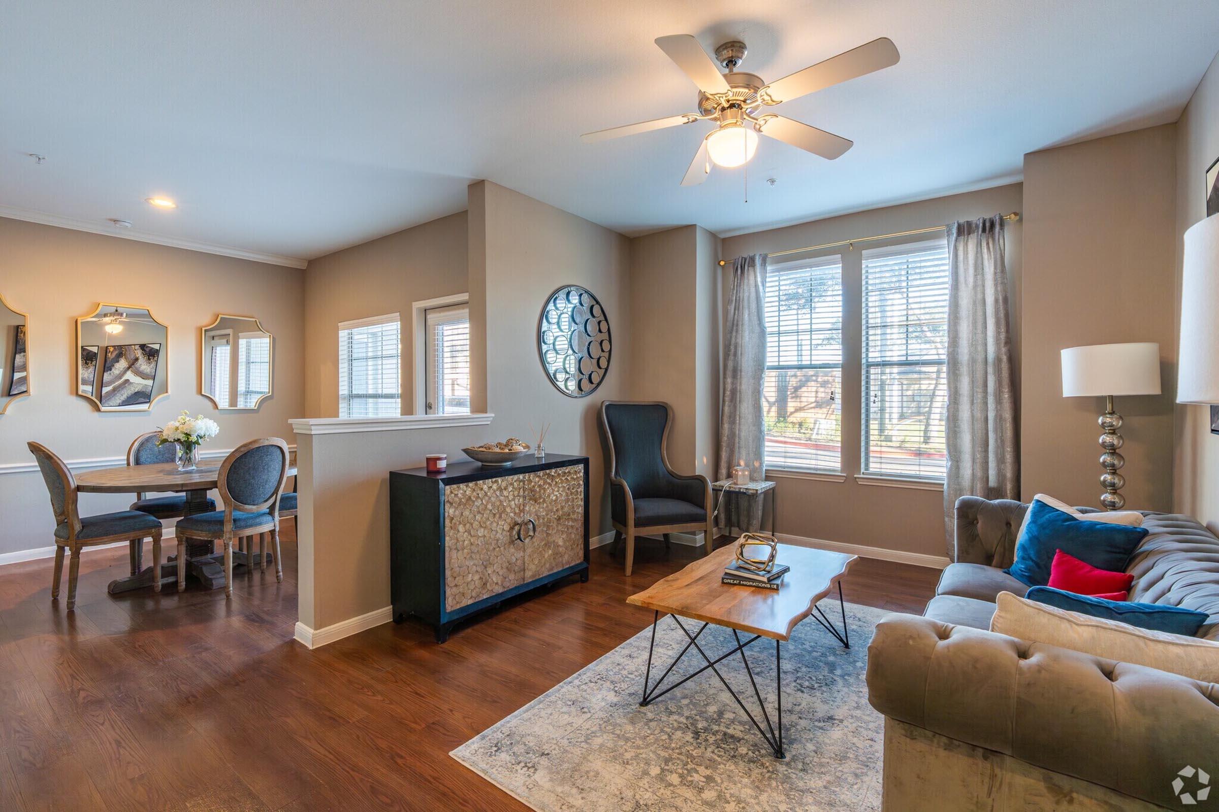A stylish living room with a beige color scheme, featuring a comfortable gray sofa, a wooden coffee table, and a decorative black cabinet. There is a ceiling fan, large windows with blinds, and two framed mirrors on the walls. A dining area with a round table and upholstered chairs is visible in the background.