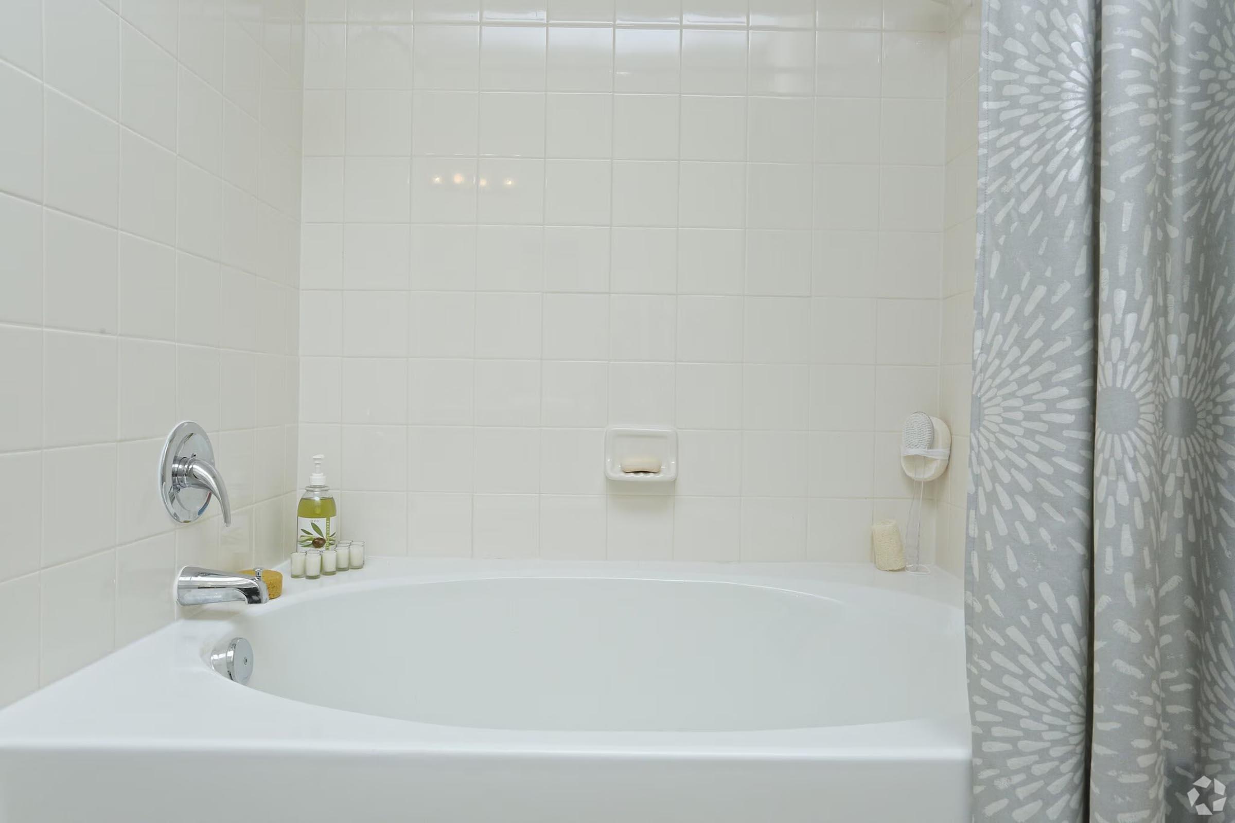 A clean, modern bathroom featuring a spacious white bathtub. The walls are tiled in light colors with a decorative shower curtain in shades of gray. Shelf items include toiletries and a bottle of bath product, creating a serene and inviting atmosphere.