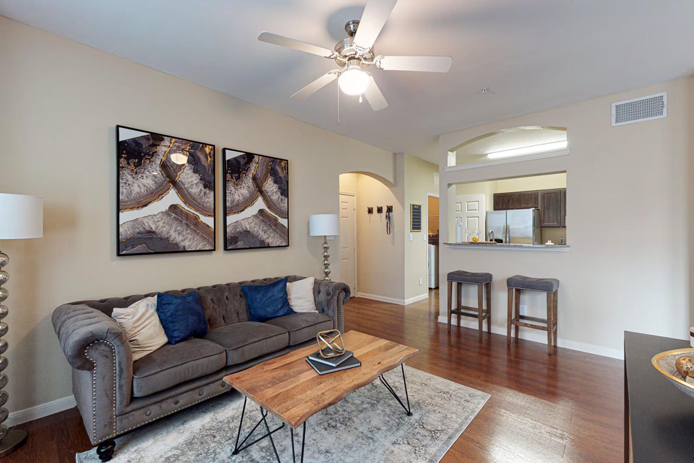 Cozy living room featuring a gray sofa with blue and white accent pillows, a wooden coffee table, and two bar stools. Wall art with abstract designs complements the warm, neutral walls. A ceiling fan hangs above, and the space leads to an open kitchen view in the background, creating a welcoming atmosphere.