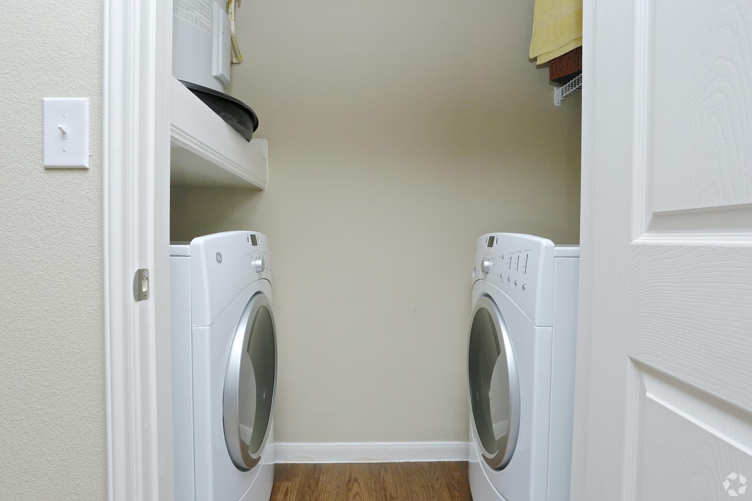 A laundry closet featuring a stacked washer and dryer side by side. The walls are painted light beige, and a shelf above holds laundry supplies. A towel hangs on a rack to the right, and the floor is made of hardwood. The space is organized and well-lit.