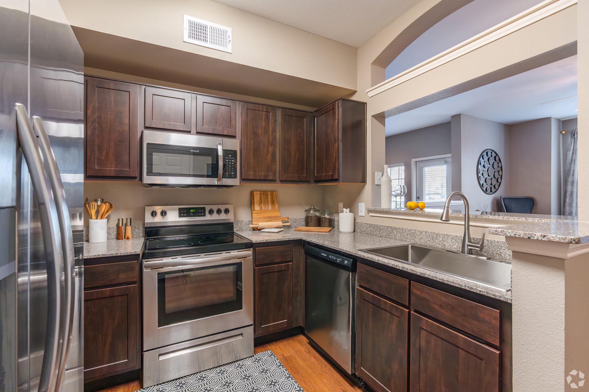 Modern kitchen featuring dark wood cabinetry, stainless steel appliances including a microwave, oven, and refrigerator, granite countertops, and a double sink. A small window adds natural light, and decorative items are displayed on the countertop. The space is tidy and contemporary, with a stylish rug on the floor.