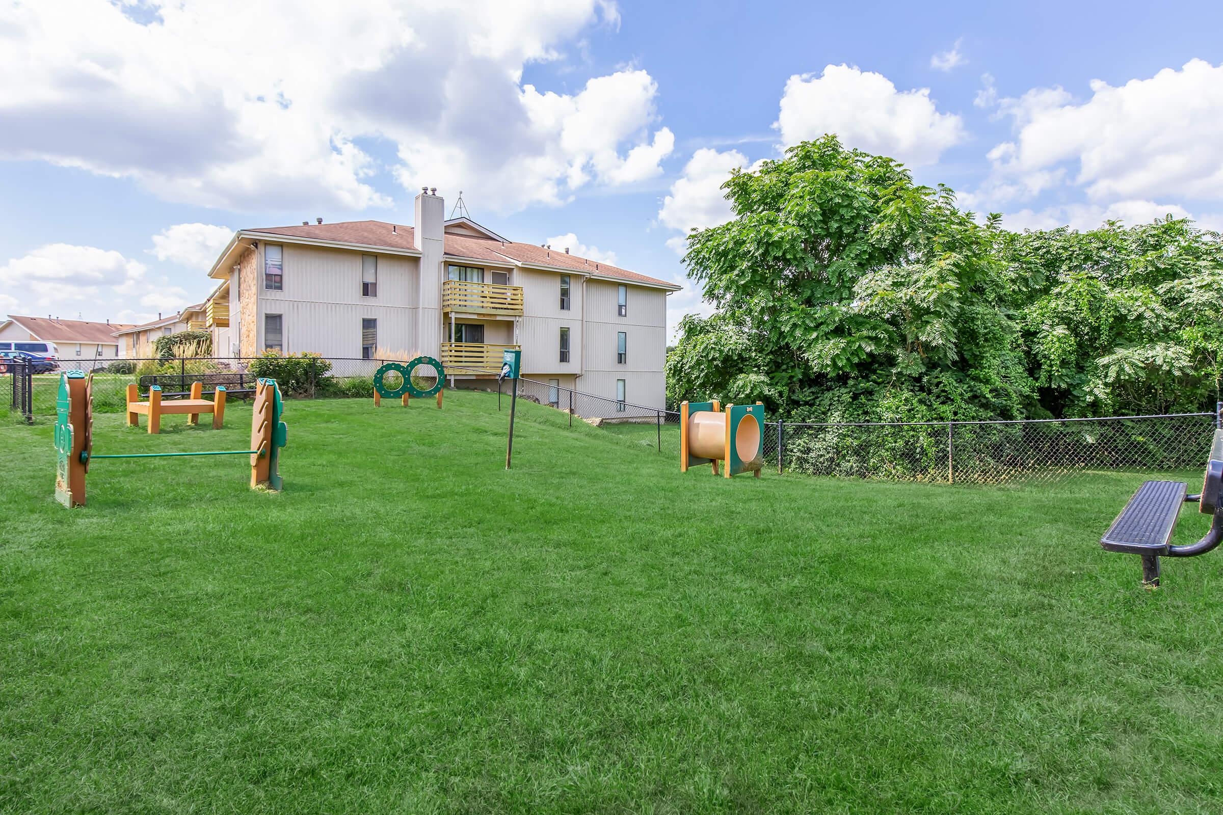A spacious green yard with playground equipment, including a tunnel, hoops, and climbing structures. In the background, there is a two-story building with a balcony, surrounded by trees and a fence. The sky is partly cloudy, adding a bright atmosphere to the scene.