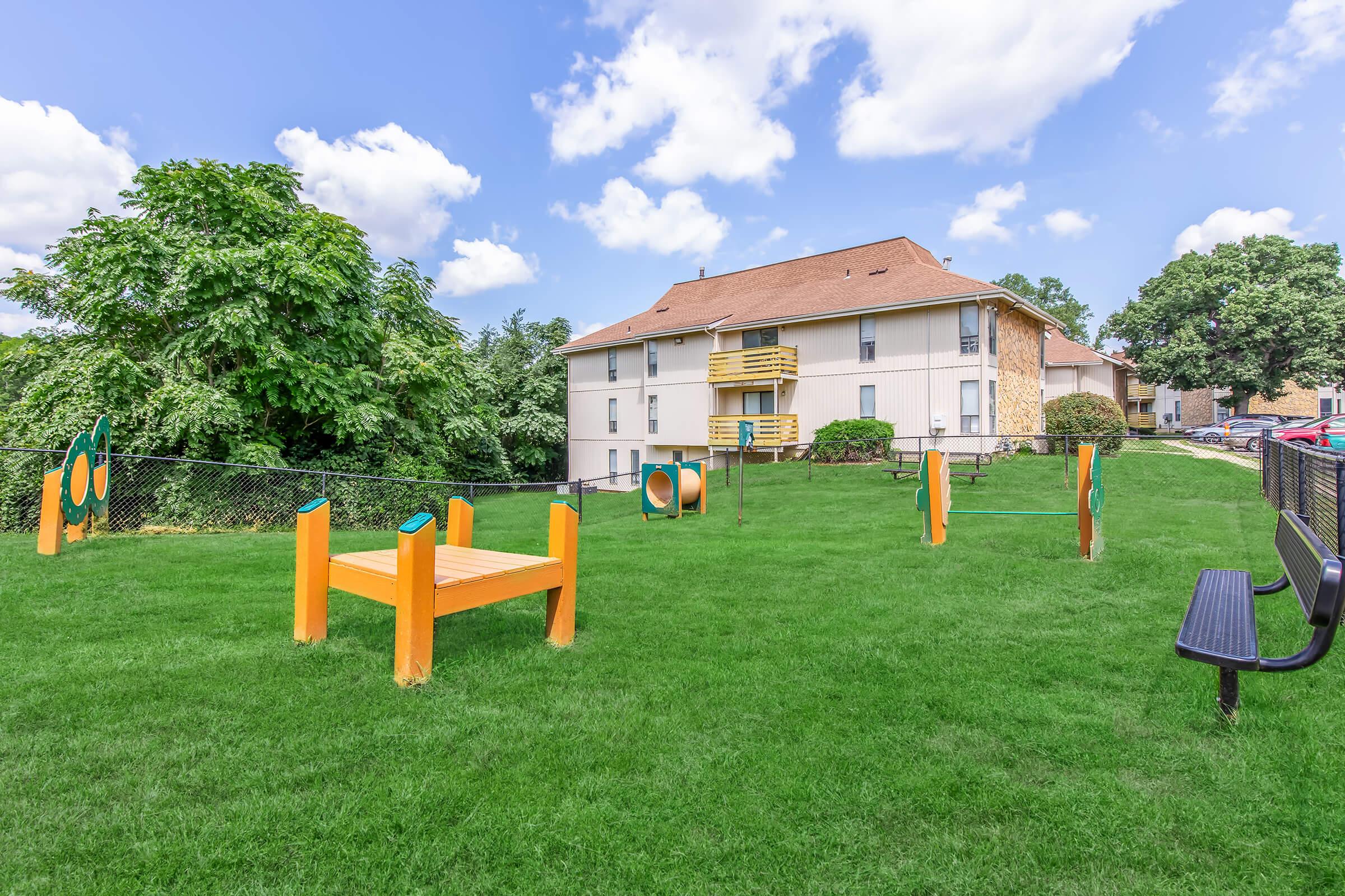 A grassy area with playground equipment, including a climbing structure and activity panels. In the background, there is a two-story building with balconies. A black bench is visible near the edge of the play area, surrounded by a fence and several trees under a blue sky with scattered clouds.