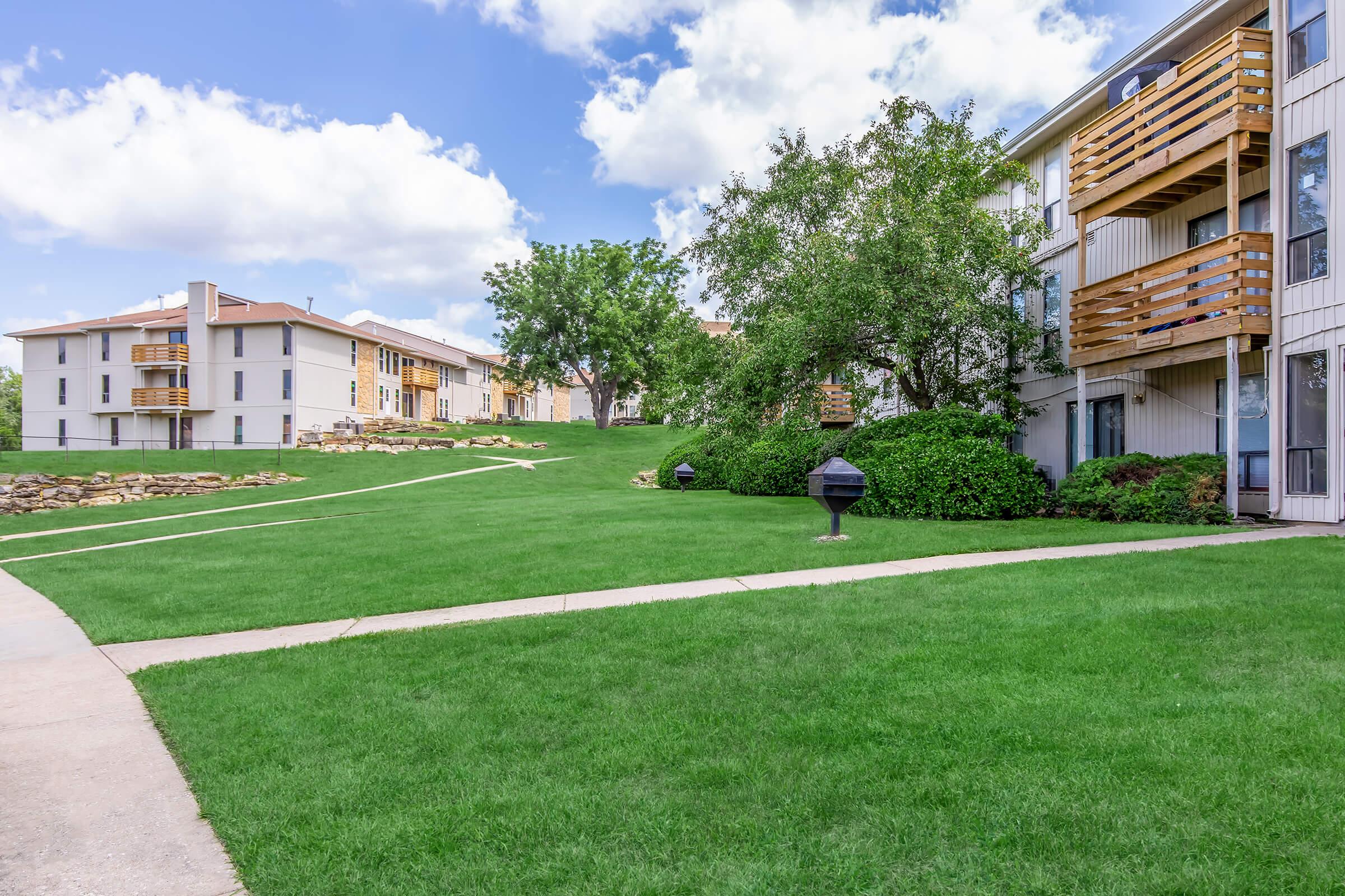 Lush green lawn with a winding pathway, accompanied by two buildings in the background. One building features balconies, while the other has a stone landscape. Several trees and barbecue grills are present, underscoring a serene outdoor setting on a sunny day with scattered clouds.