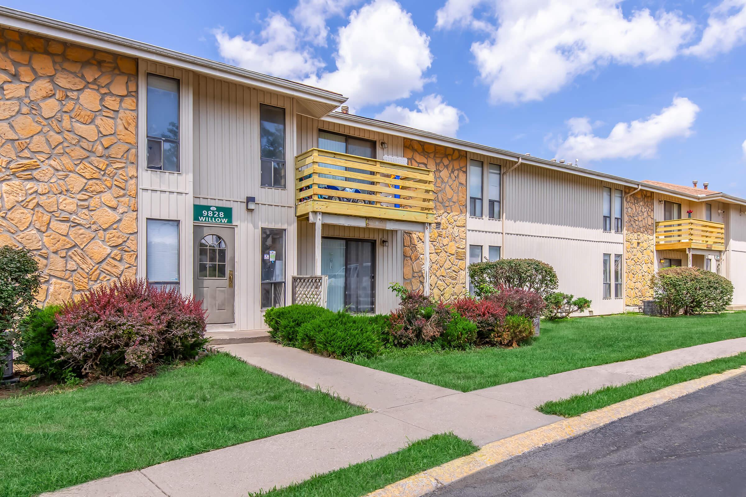 Two-story apartment building with a stone façade and beige siding. Each unit features a yellow balcony railing. Lush green grass and shrubs line the walkway leading to the entrance, under a partly cloudy blue sky.