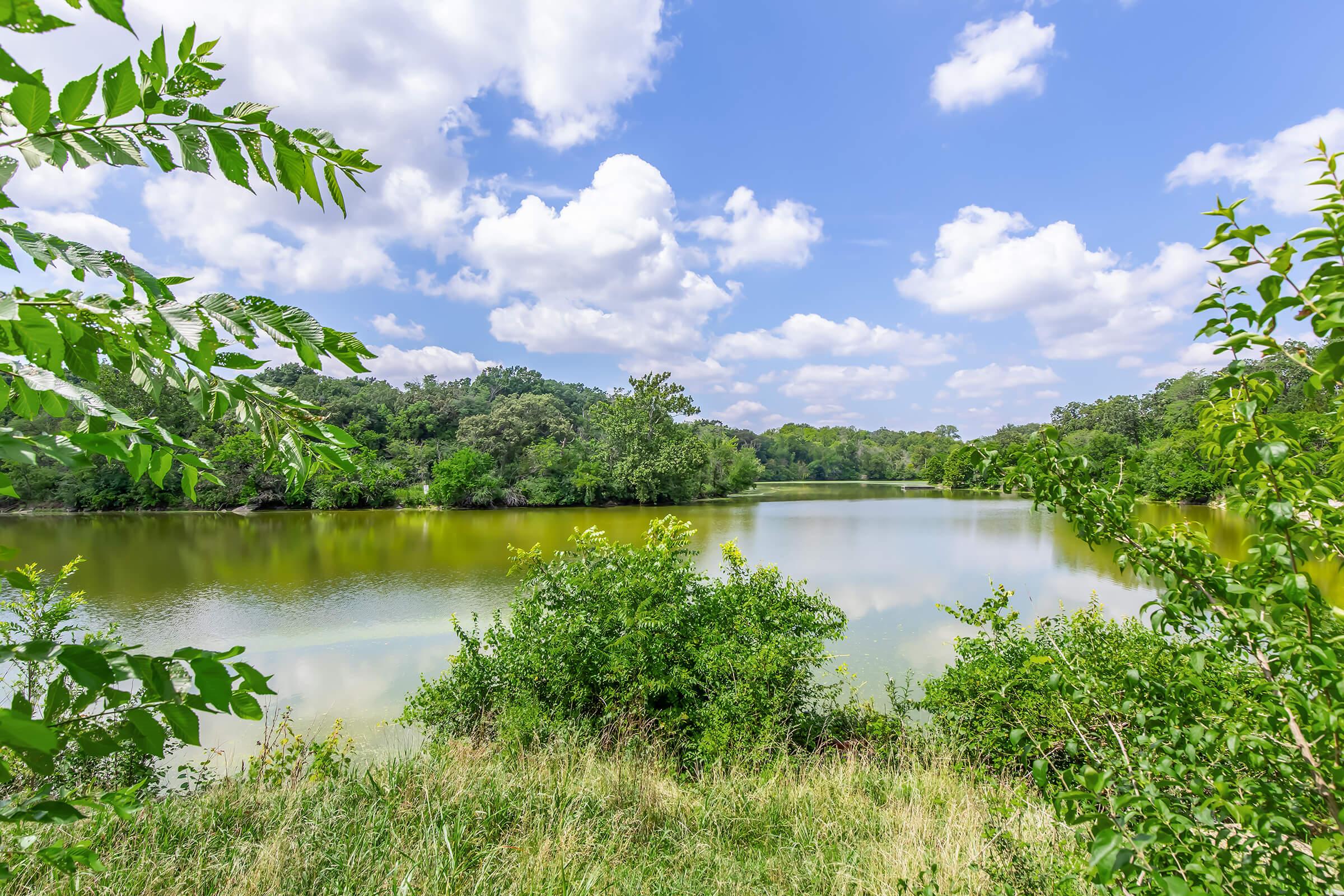 A serene landscape featuring a calm river surrounded by lush greenery, trees lining the banks, and a bright blue sky with fluffy white clouds. The water reflects the natural beauty of the surroundings, creating a peaceful and idyllic outdoor scene.