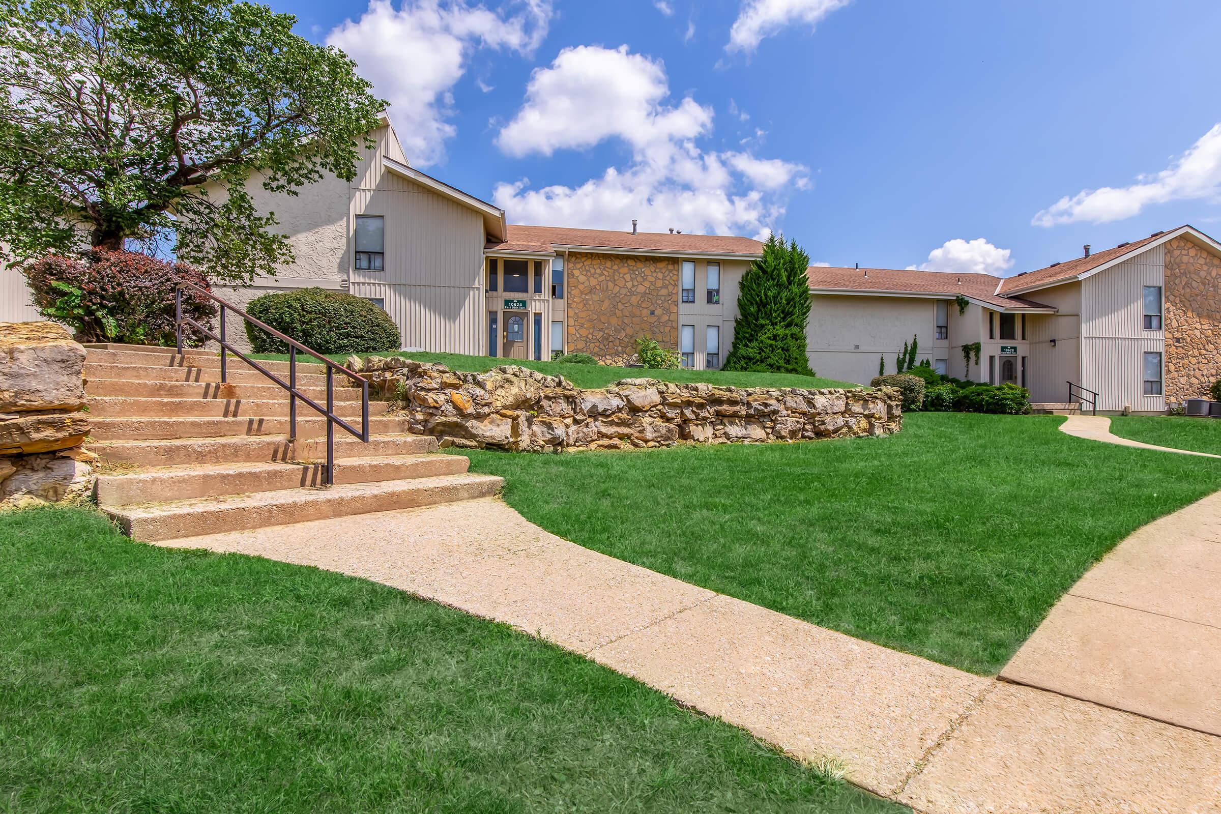 A landscaped outdoor area featuring two multi-story apartment buildings. There are well-manicured lawns with vibrant green grass, stone steps leading up to the buildings, and a rocky border. The sky is bright blue with fluffy white clouds.