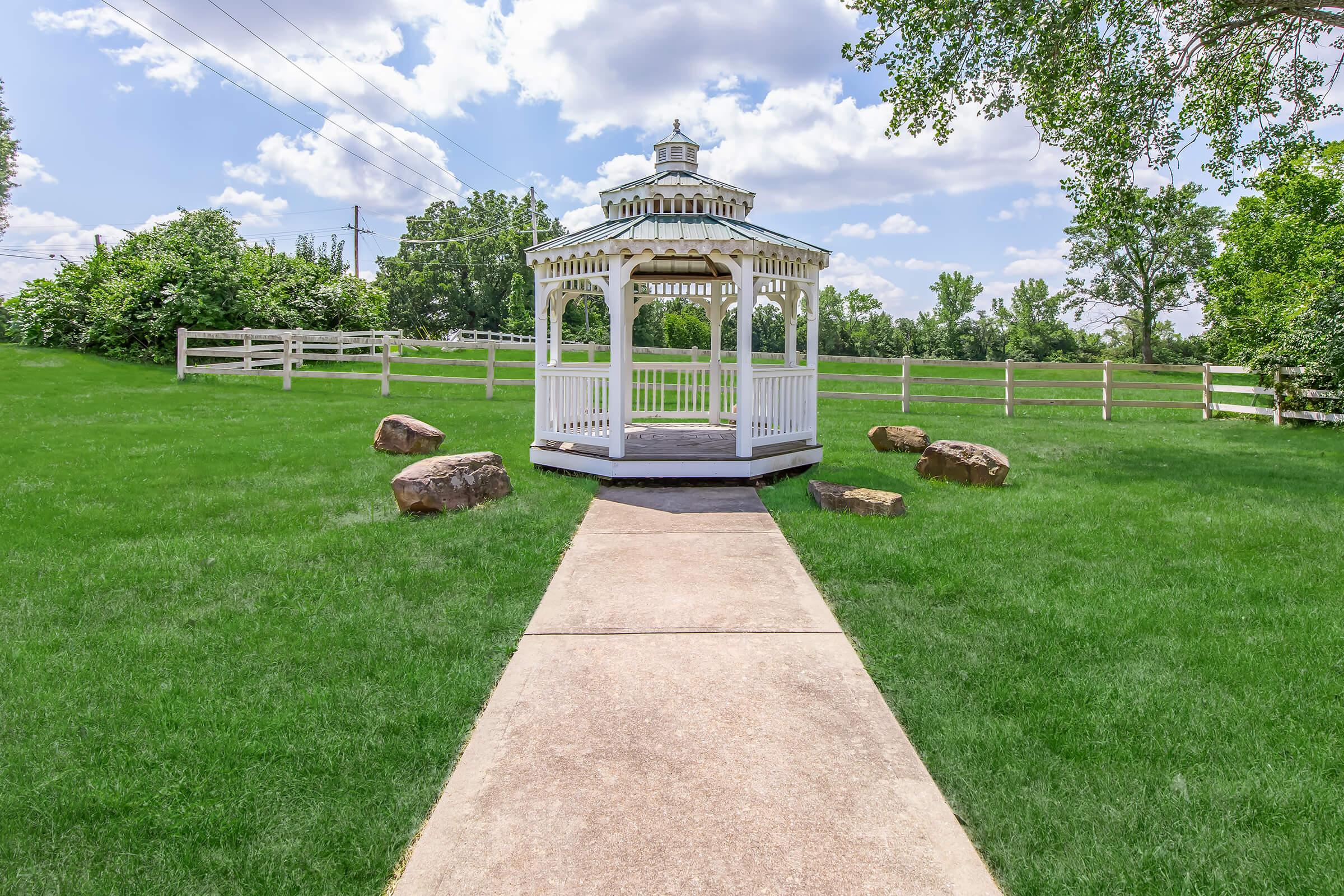 A white gazebo with a peaked roof sits at the end of a concrete pathway, surrounded by lush green grass. Large stones are placed along the path, and trees and a white fence are visible in the background under a partly cloudy sky.