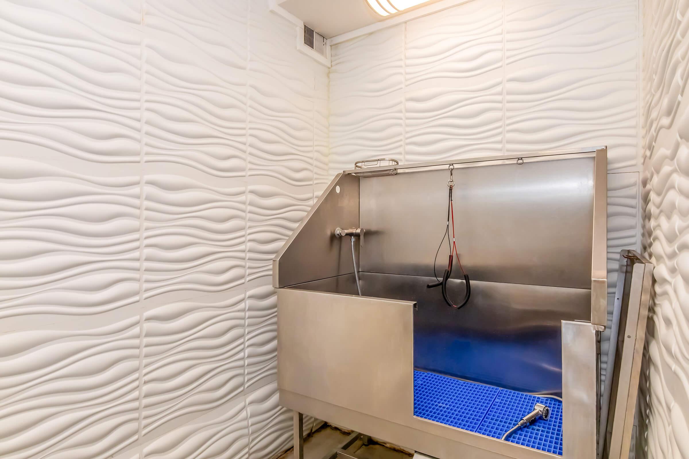 A stainless steel tub with a faucet and hoses, set against a textured, wavy white wall. The tub features a blue grid base and is designed for washing or rinsing. The overall setting is simple and utilitarian, suggesting a space for maintenance or grooming activities.