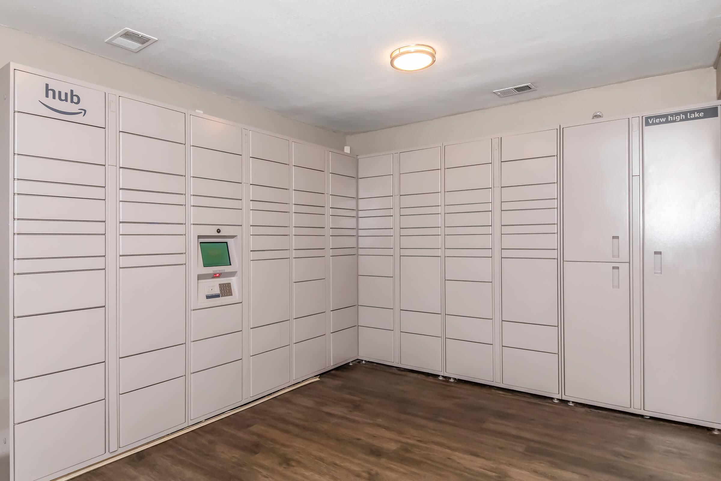 A clean and organized room featuring Amazon hub lockers for package delivery. The lockers are light-colored, arranged neatly against the walls, with an interactive touchscreen kiosk in the center. The flooring is wooden, and there are no visible windows, creating a functional, secure space for parcel collection.