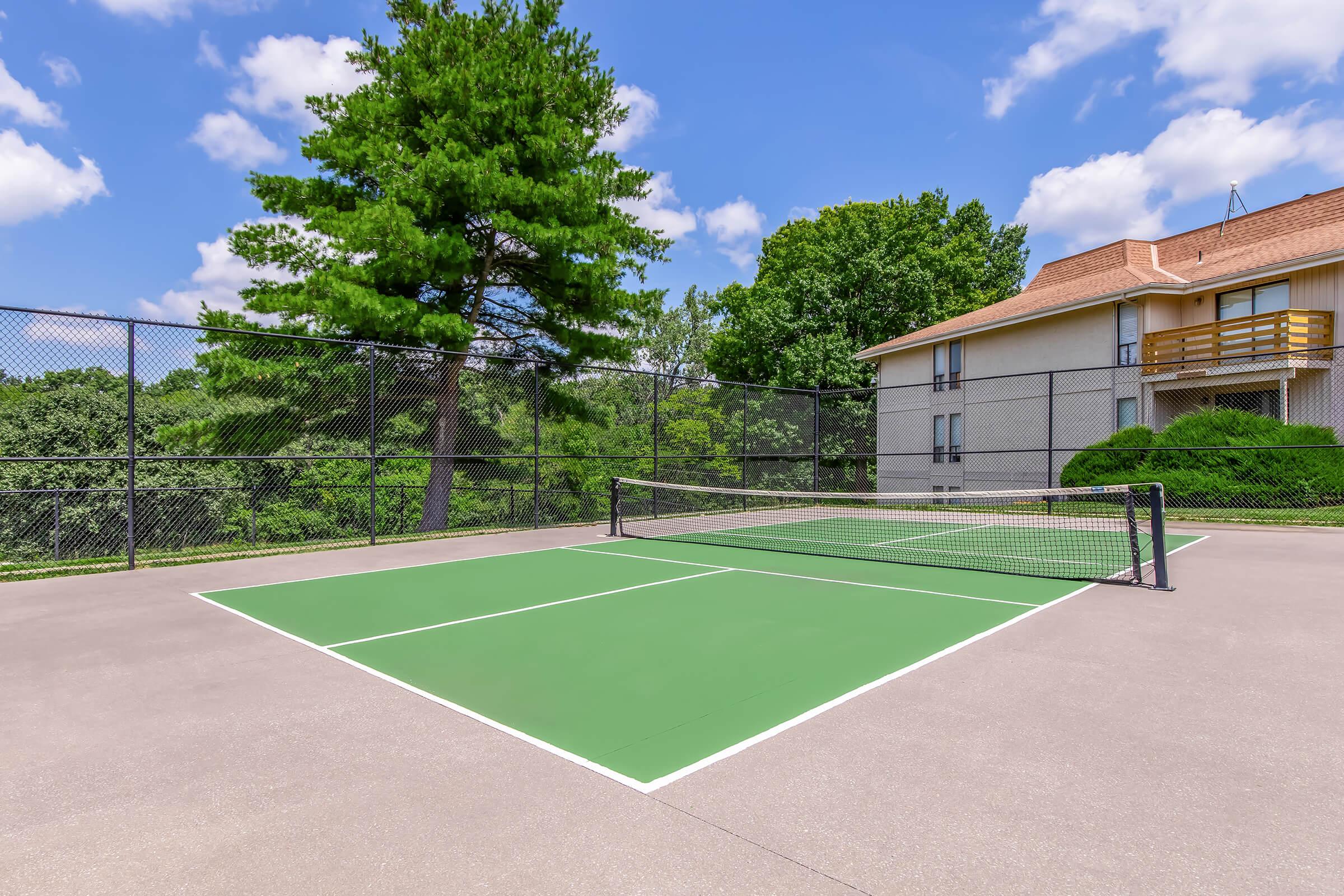 A tennis court featuring a green playing surface, surrounded by a black chain-link fence. In the background, there are trees and a residential building with balconies. The sky is clear with a few clouds, creating a bright and inviting outdoor space.