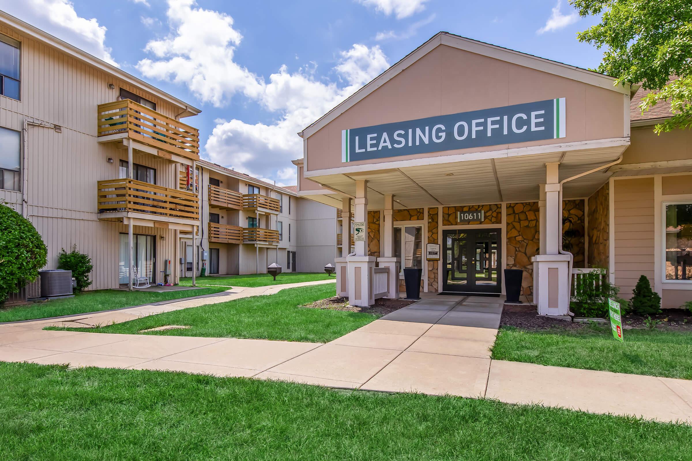 Leasing office entrance of an apartment complex, featuring a welcoming porch with a sign, surrounded by grassy areas and light-colored buildings. Cloudy blue sky in the background adds to the pleasant atmosphere.