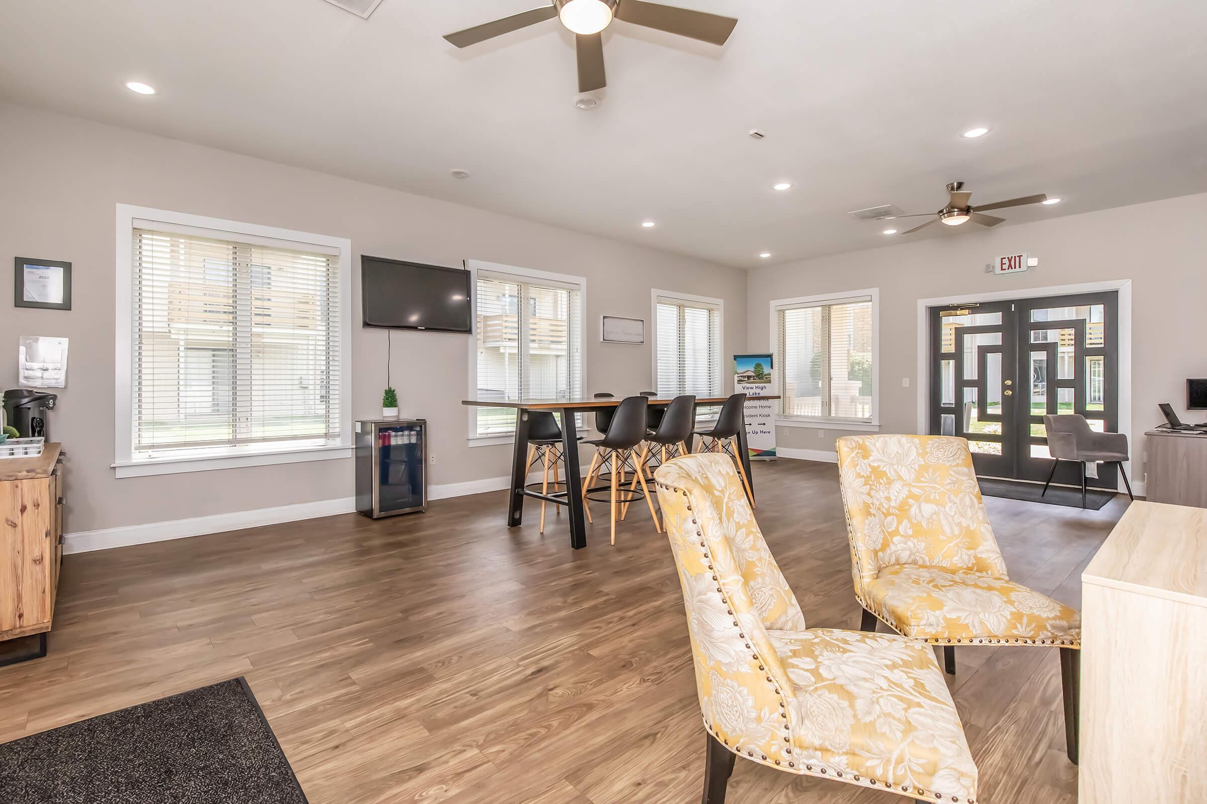 Bright and modern community lounge featuring a wooden floor, two patterned chairs, and a large window. There's a round table with four chairs, a TV on the wall, and a kitchenette area. Natural light fills the space, creating a welcoming atmosphere. Ceiling fan visible above.