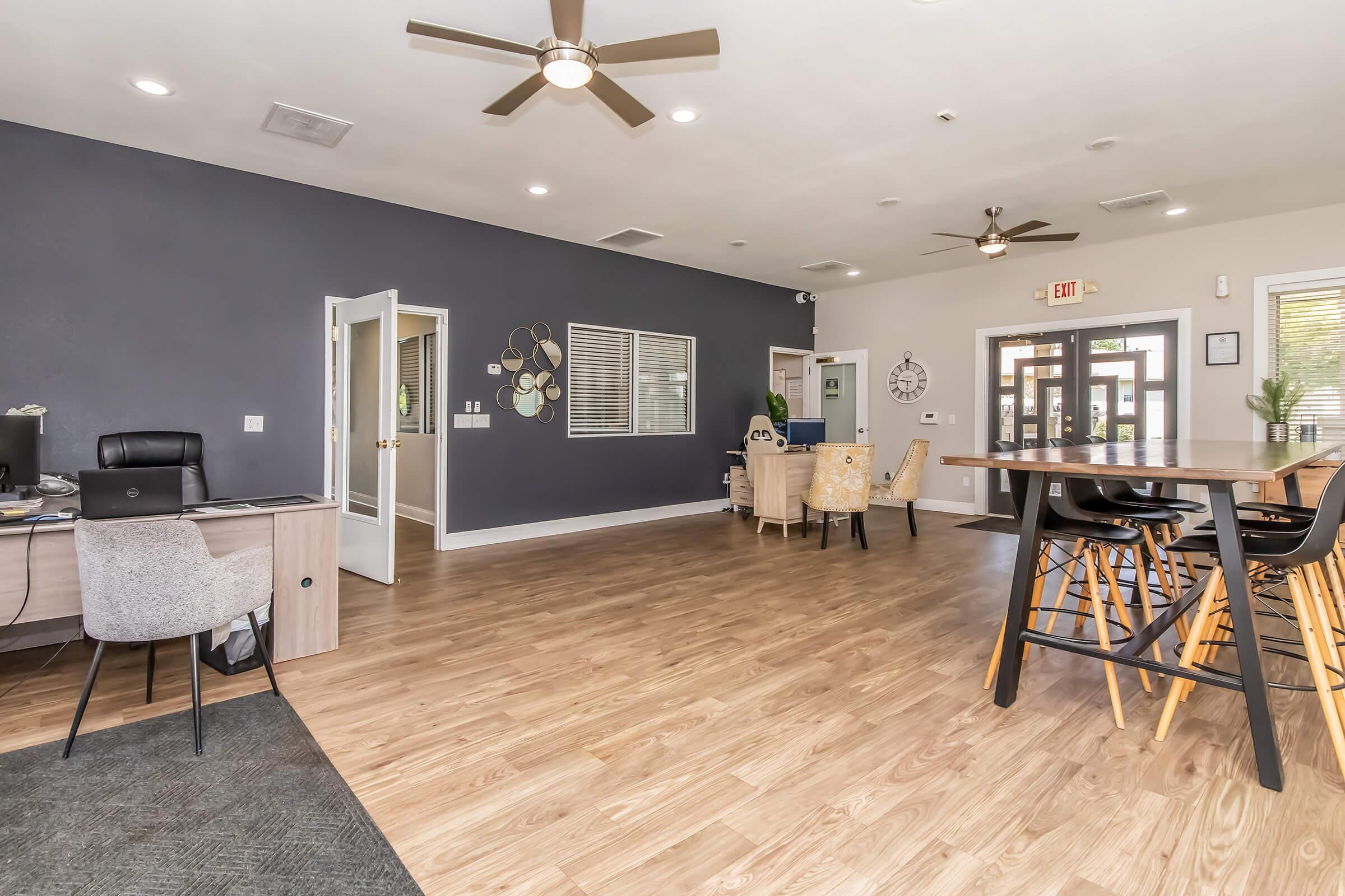 Interior view of a modern communal space featuring a light wood floor, a desk area with a black chair, and a dining area with elegant chairs. The walls are painted in gray tones, and there are ceiling fans and large windows for natural light. Decor includes wall art and a welcoming entrance.