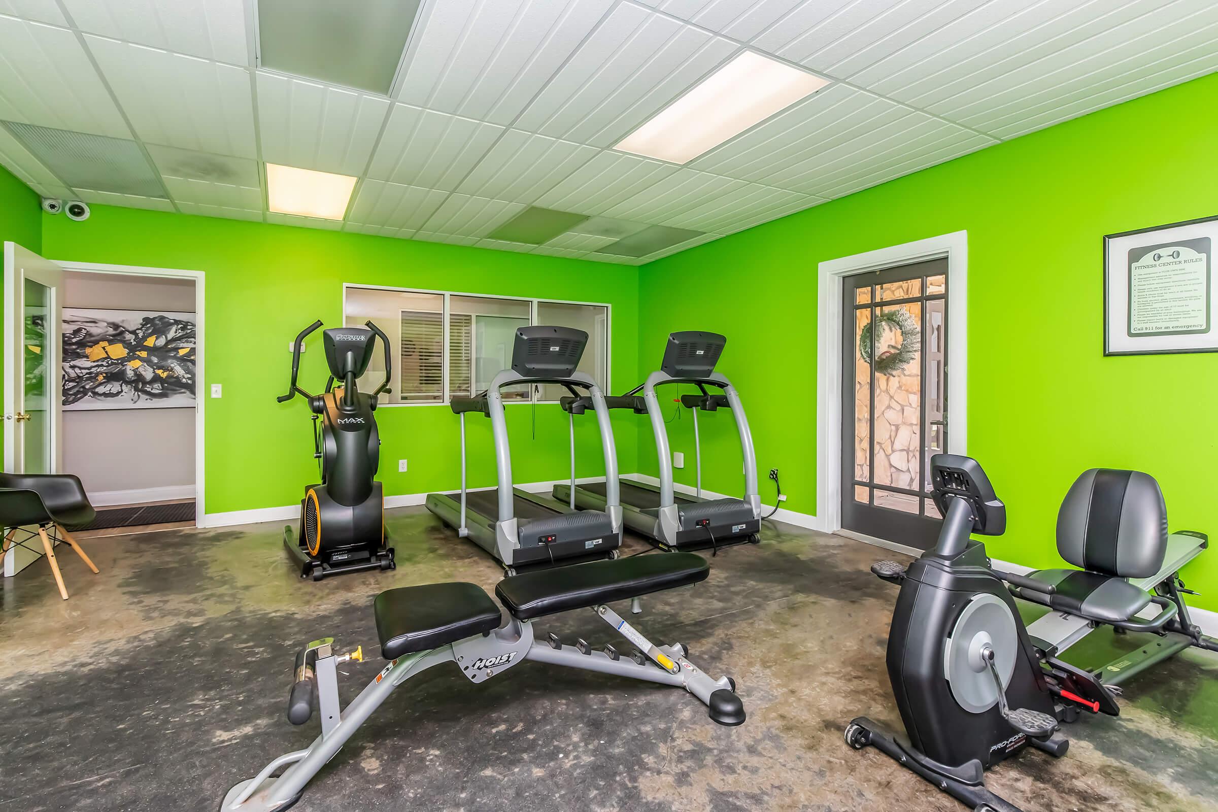 A brightly colored gym interior featuring green walls, equipped with exercise machines including two treadmills, a stationary bike, and a weight bench. The space is well-lit, with a window and a door leading outside, creating an inviting workout environment.