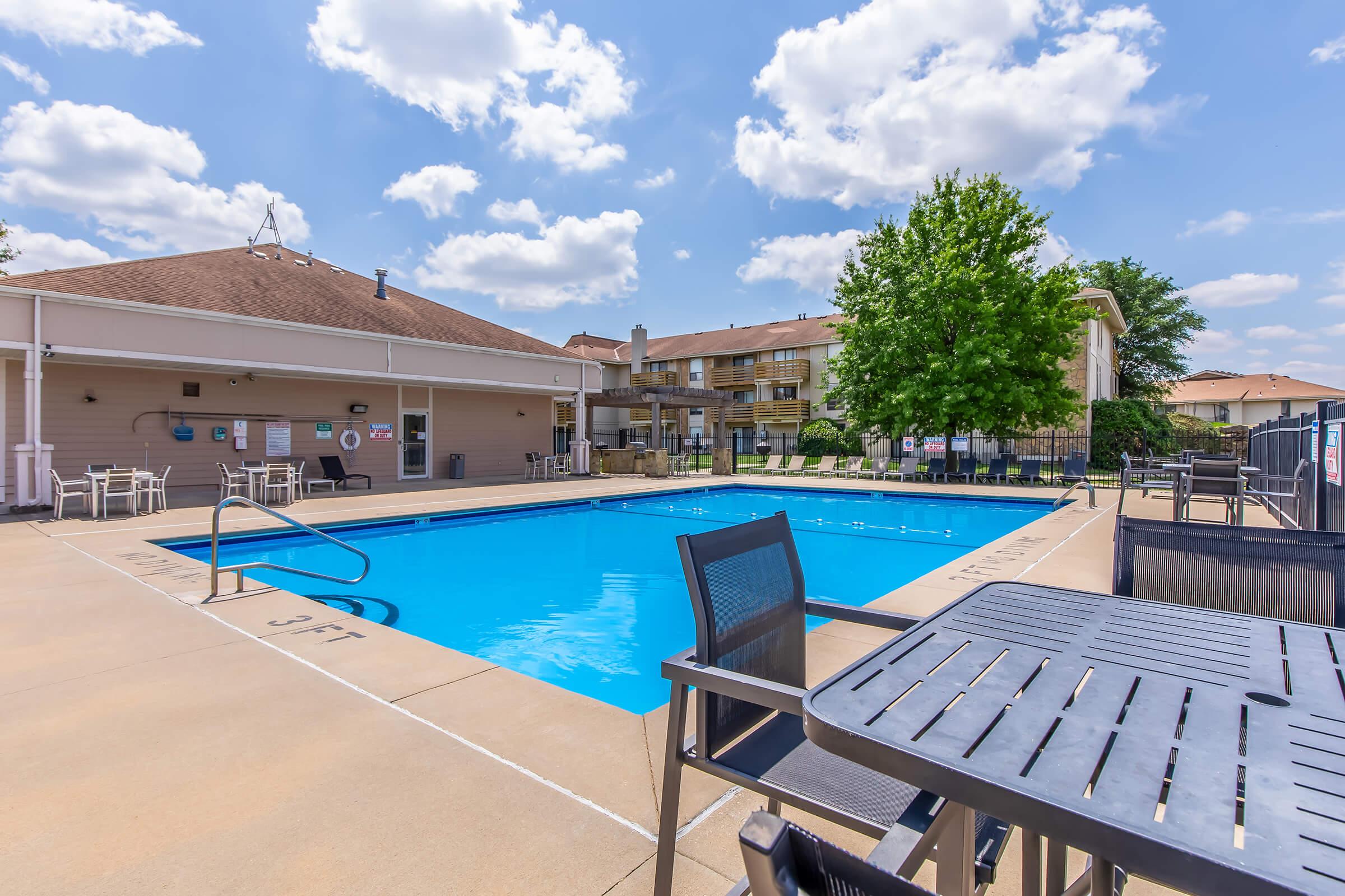 A clear blue swimming pool surrounded by lounge chairs and tables, with a building in the background. The sky is partly cloudy, and there are trees nearby, creating a relaxing outdoor atmosphere. Ideal for leisure and recreation.