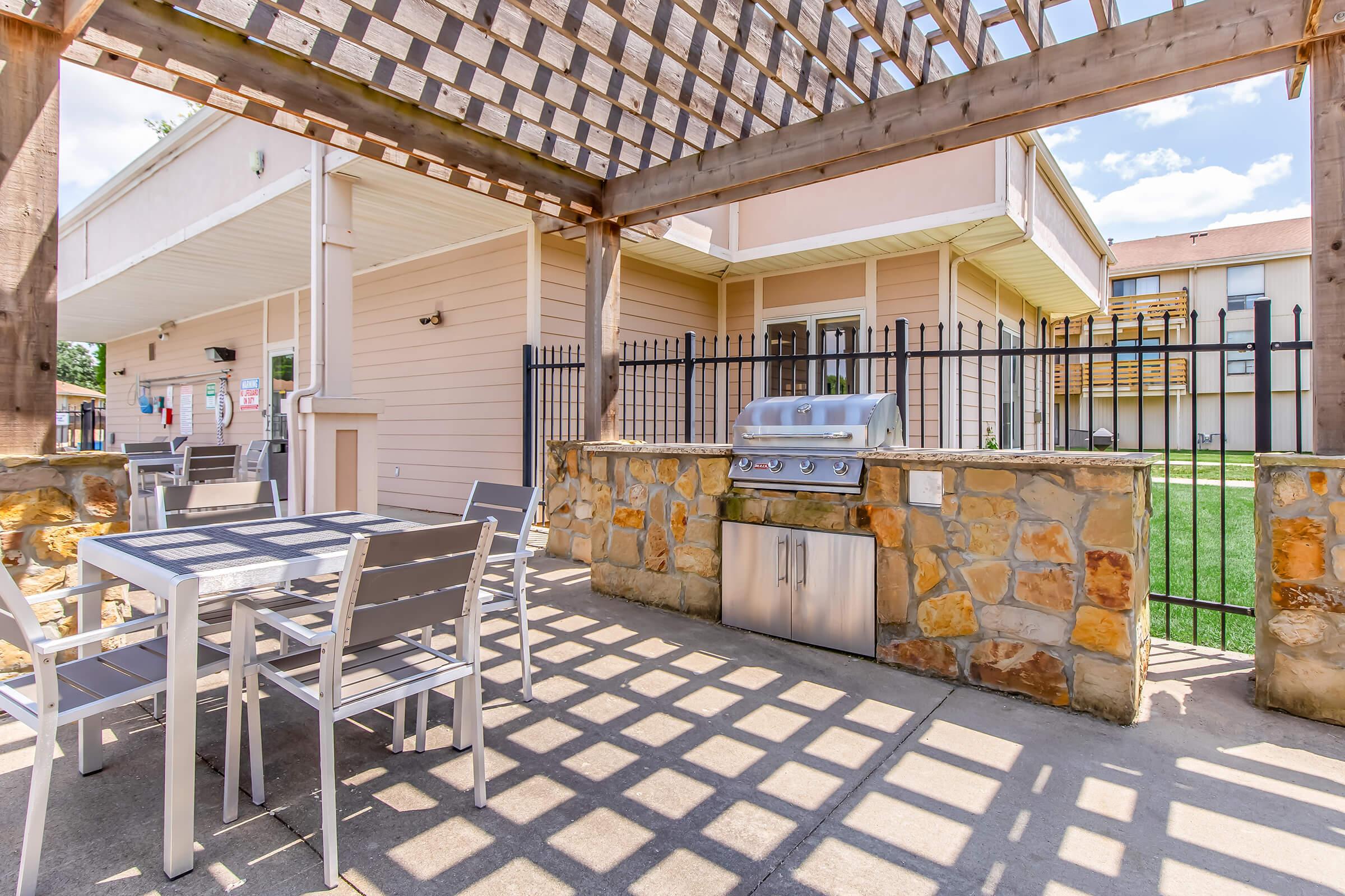 Outdoor grilling area featuring a stainless steel barbeque grill beneath a wooden pergola, surrounded by a stone wall. There are metal outdoor tables and chairs set up for dining. Green grass and buildings are visible in the background under a clear blue sky.