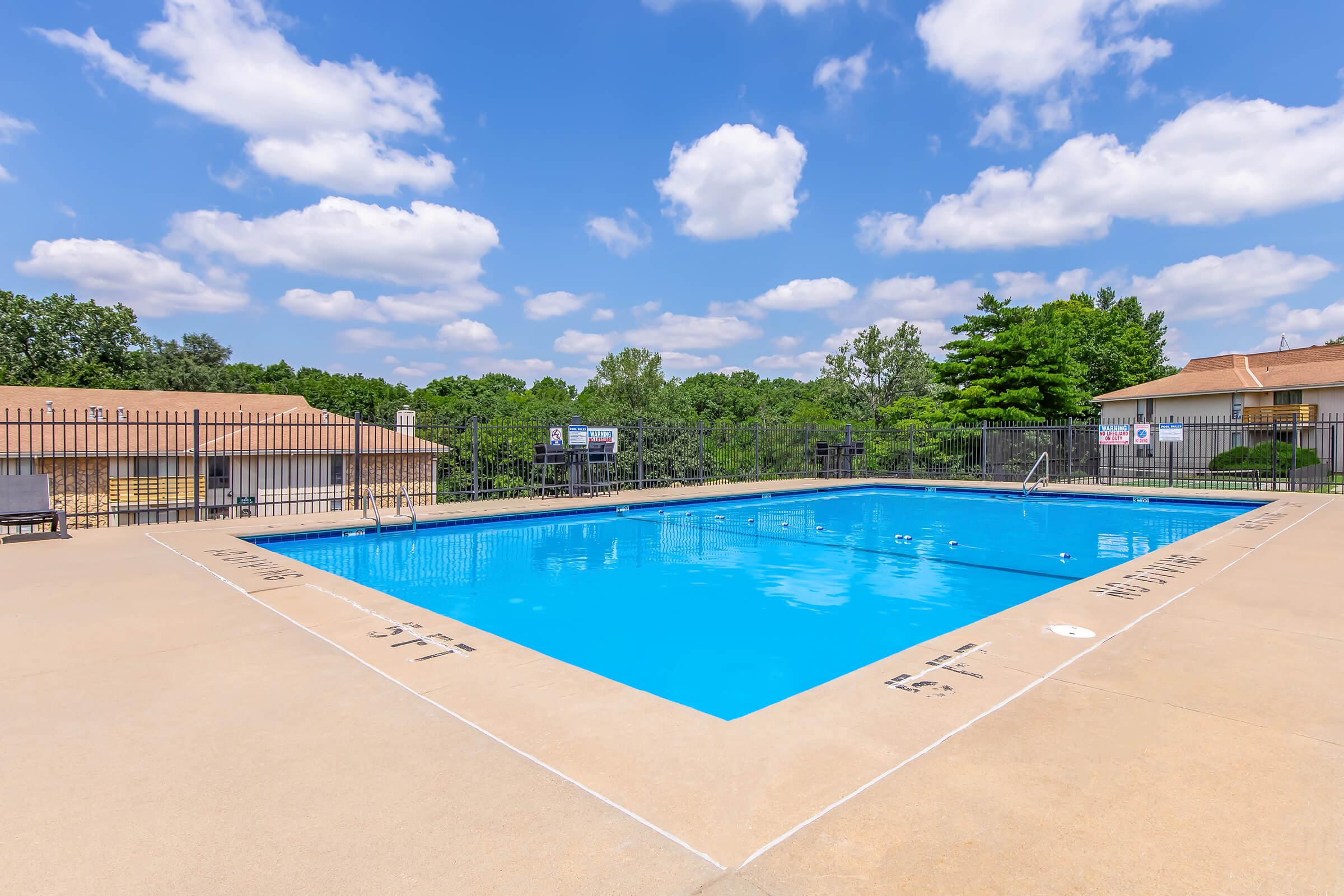 A clear blue swimming pool surrounded by a concrete deck and black metal fence. The scene includes a few trees in the background under a bright blue sky with fluffy white clouds. There are no people visible in the area, creating a tranquil atmosphere.