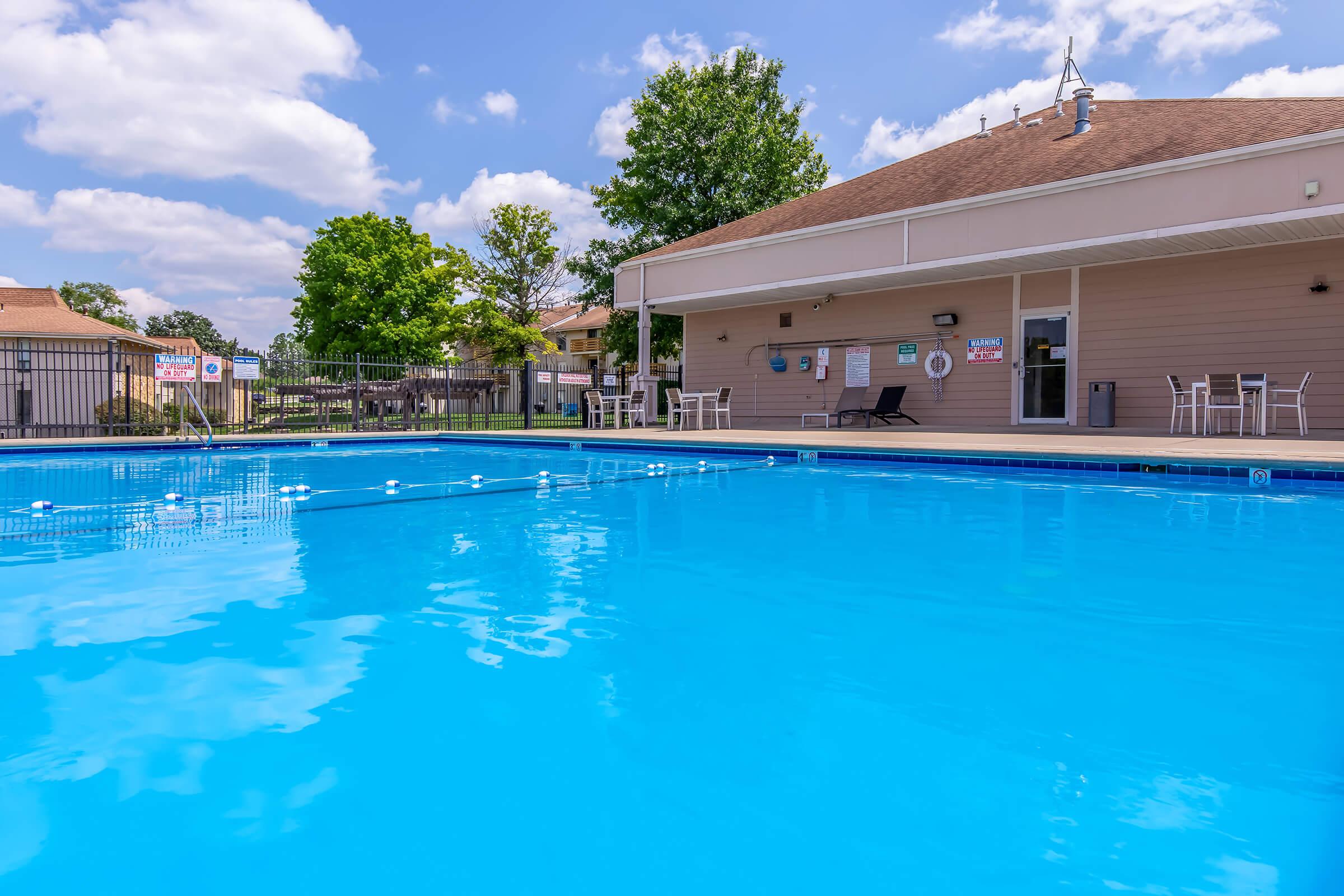 A clear blue swimming pool with lounge chairs and tables nearby. The pool area is surrounded by trees and a fence, under a bright sky with some clouds. A building with large windows and entrance is visible in the background, creating a welcoming atmosphere for relaxation and leisure.