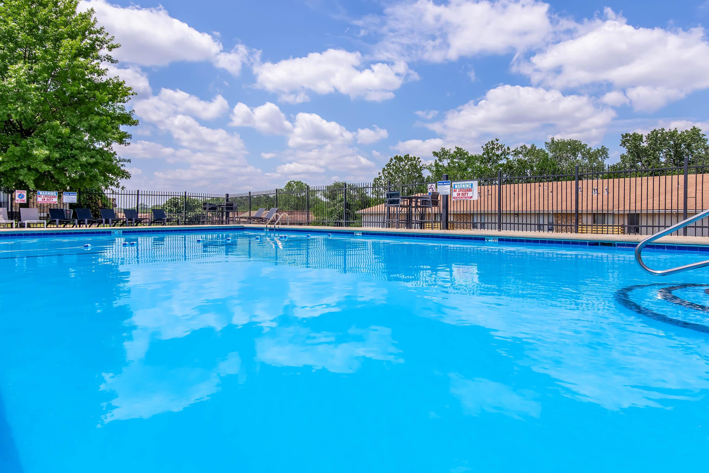 A bright blue swimming pool captured under a clear sky with fluffy clouds. The pool reflects the sky and surrounding trees. In the background, there are lounge chairs and a fence, adding to a serene outdoor atmosphere. Ideal for relaxation and swimming.