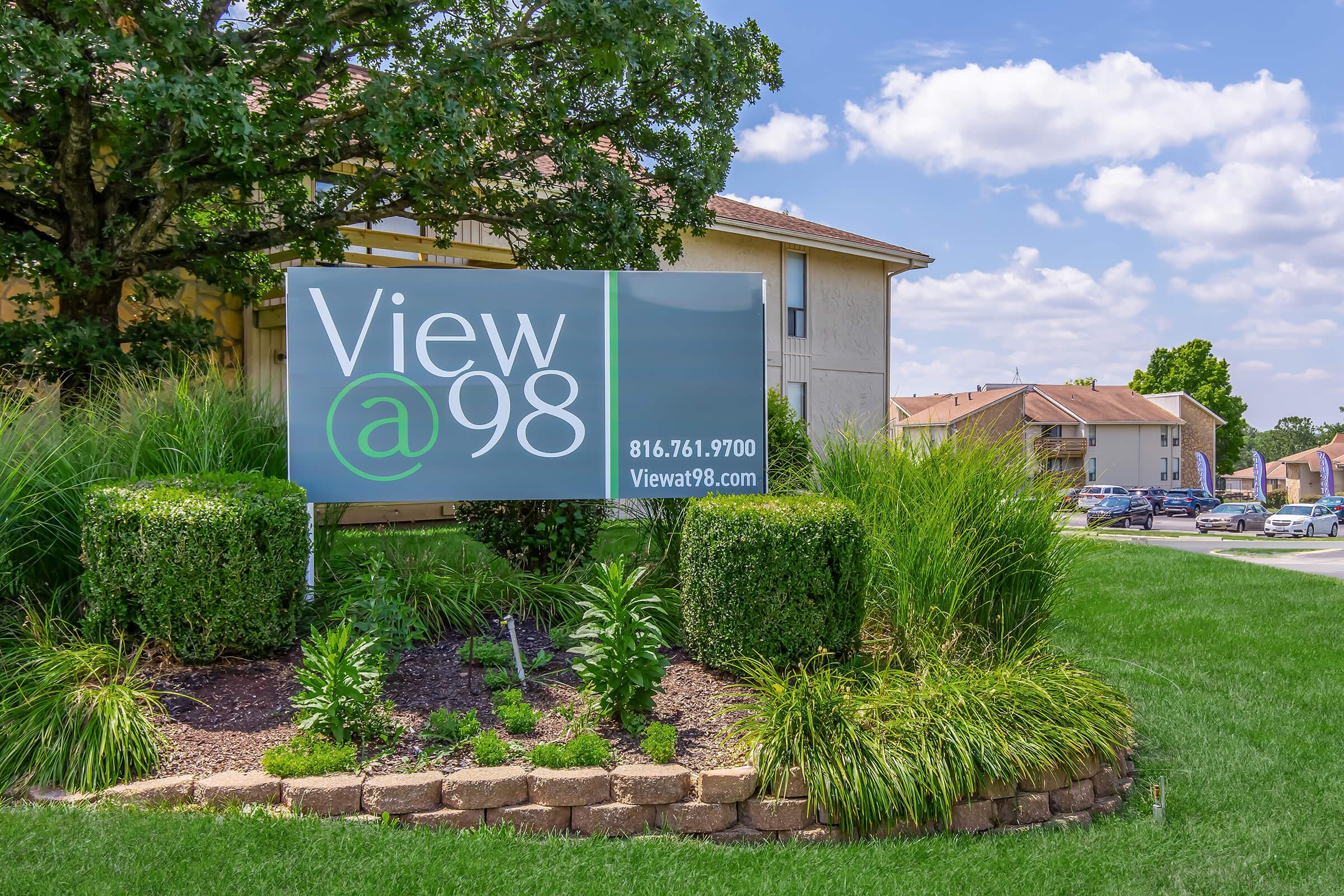Sign for "View @ 98" featuring contact information (816.761.9700) and website (Viewat98.com), surrounded by landscaped greenery and residential buildings in the background under a partly cloudy sky.