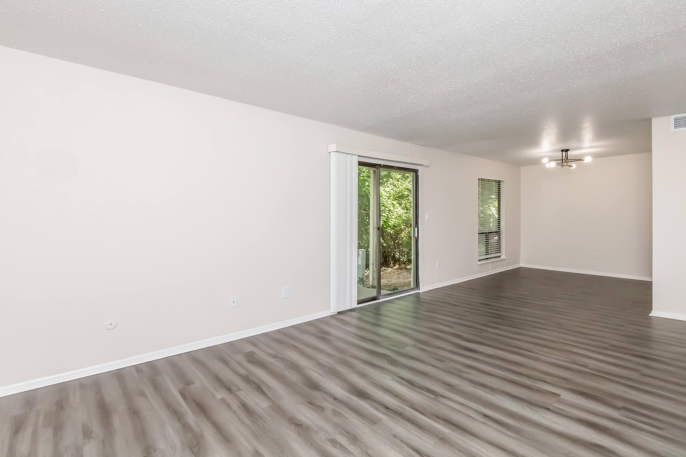 Empty living room with light-colored walls and wood-style flooring. A sliding glass door leads to an outdoor area with greenery. Natural light enters the space, which features a ceiling light fixture and large windows without curtains. The room is spacious and ready for furnishings.