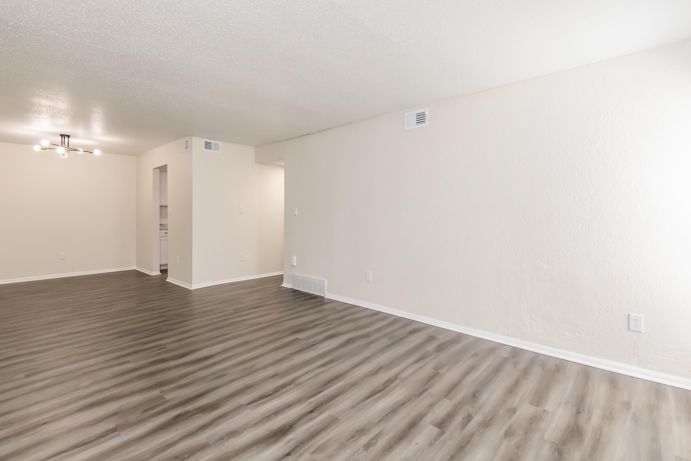A spacious, well-lit room with light-colored walls and a modern light fixture. The floor features a sleek, light wood laminate. The room has a clean, minimalist aesthetic, with no furniture, providing a blank canvas for decorating. The adjacent area shows a glimpse of a kitchen.