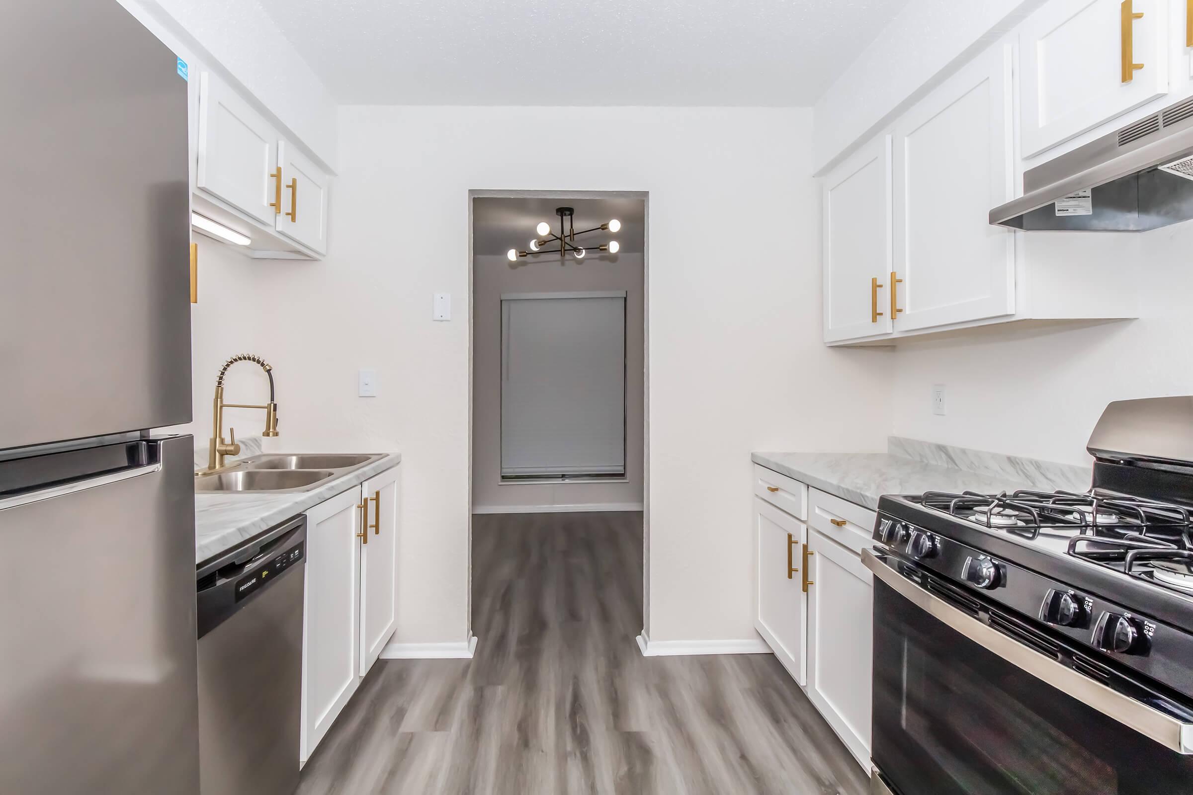 A modern kitchen featuring stainless steel appliances, including a refrigerator, dishwasher, and gas stove. The cabinets are white with gold handles, and the countertop has a marbled design. A doorway leads to a well-lit room with a large window and wooden flooring.