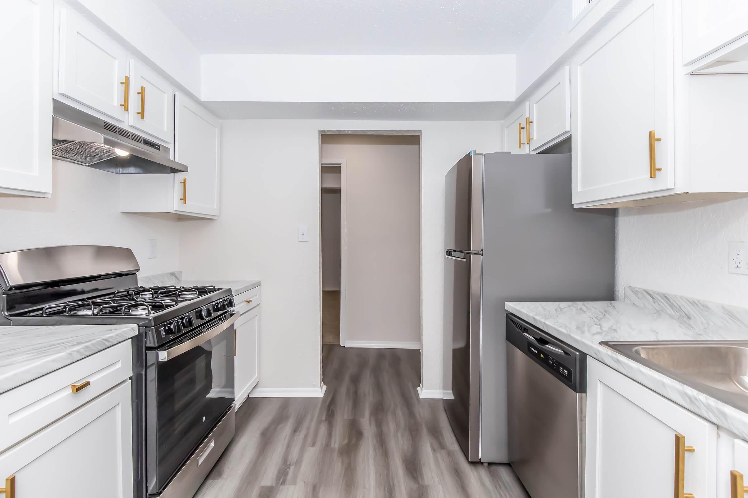 A modern kitchen featuring white cabinetry with gold handles, stainless steel appliances including a gas range, refrigerator, and dishwasher, and a marble countertop. The flooring is light gray laminate, and there's an open entryway leading to another room in the background.