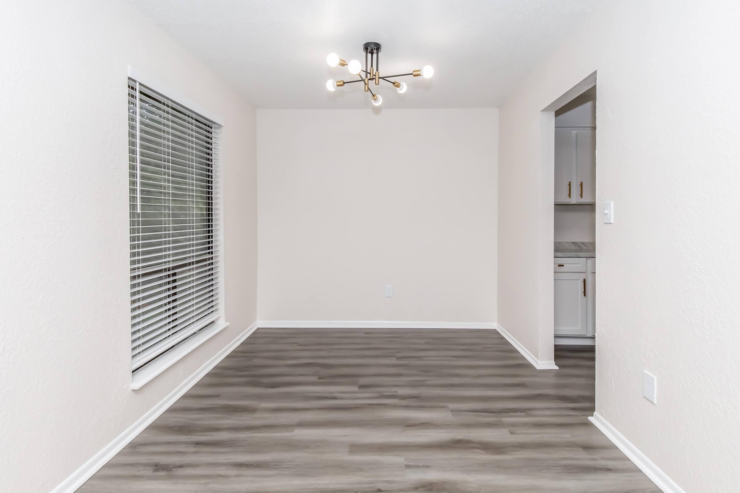A spacious, empty room with light-colored walls and wood-like flooring. A modern light fixture with multiple bulbs hangs from the ceiling. There is a large window with blinds, allowing natural light to enter, and a doorway that leads to another room with kitchen elements visible.
