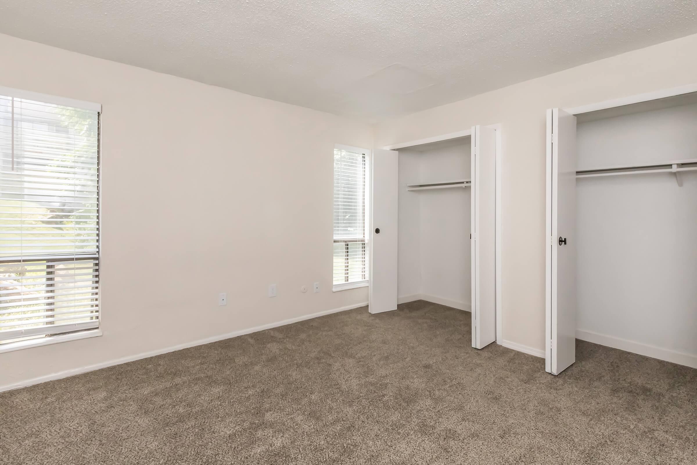 A light and airy room with beige walls and plush carpeting. Two white-paneled closets are visible, each with sliding doors and shelving inside. The room features two windows with blinds, allowing natural light to filter in. Ideal for bedroom or office space.