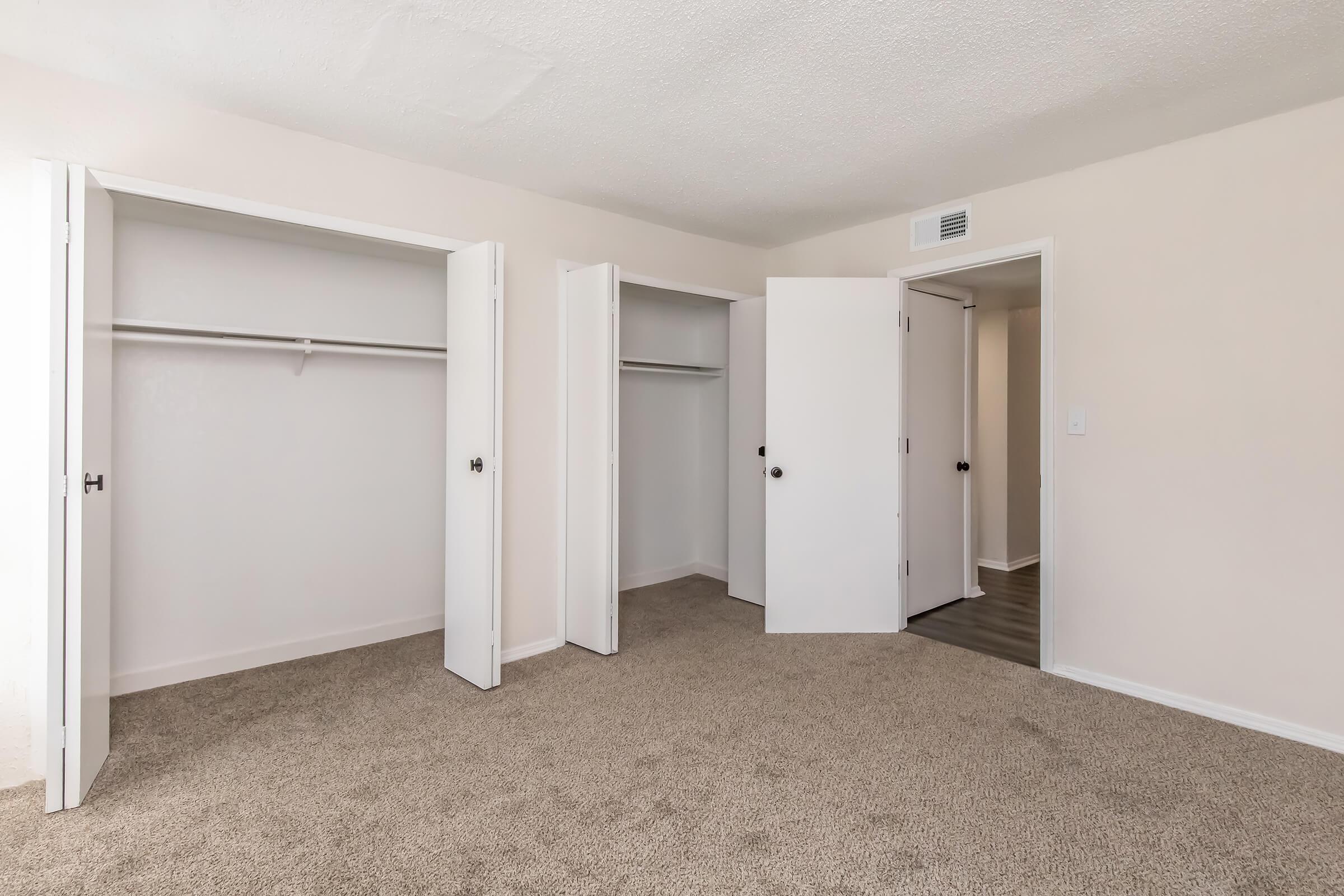 A spacious, empty room with light beige walls and tan carpeting. There are two open closet doors on the left, revealing empty shelves. A white door on the right leads to another room, and the overall space is bright and uncluttered, suitable for personal decoration.
